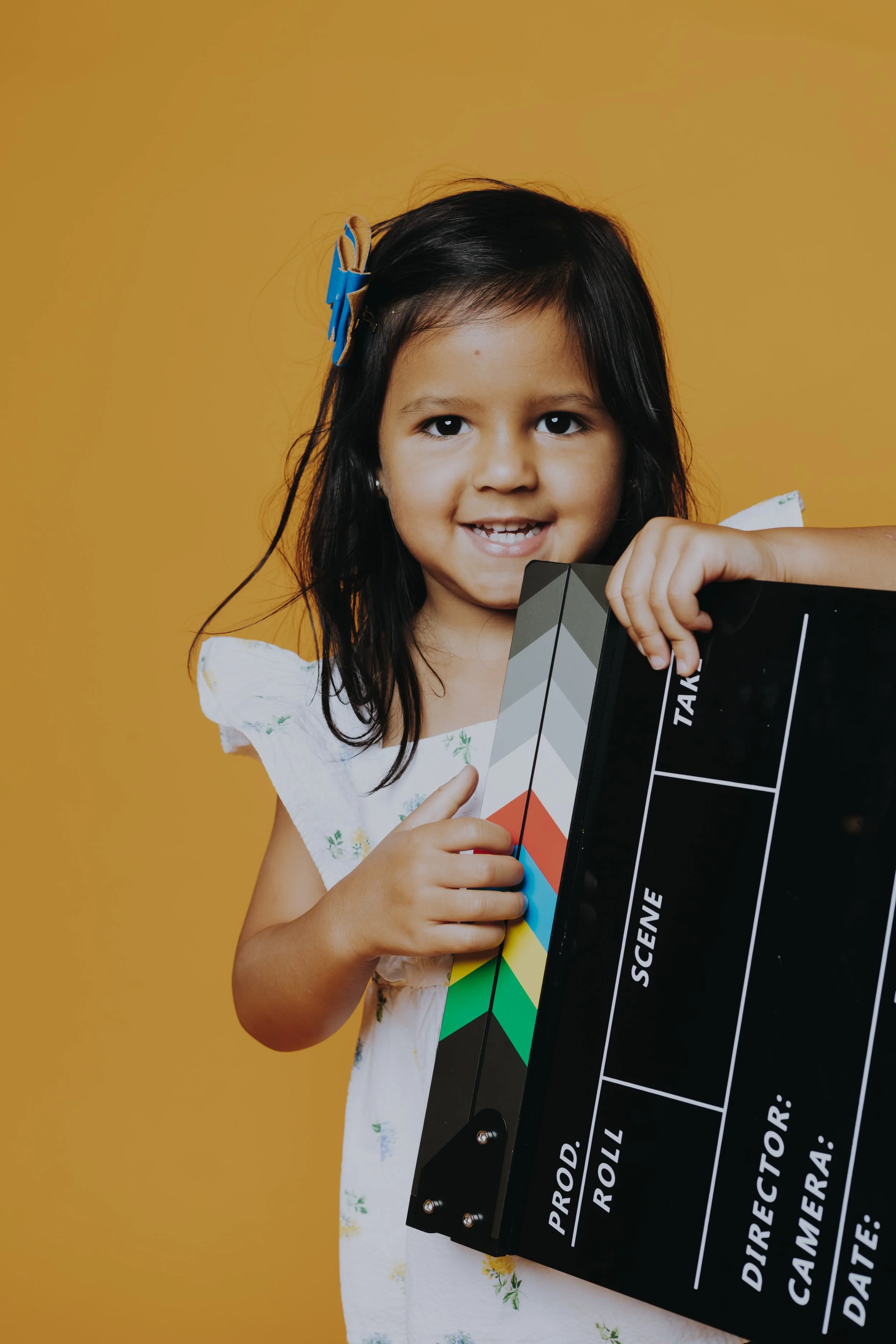 A young girl holding a movie clapperboard, smiling, against a yellow background.
