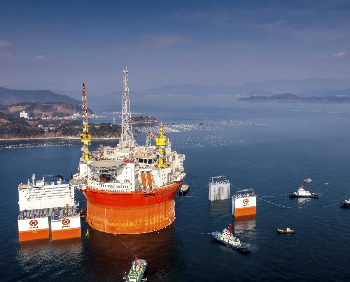 Large offshore oil or gas platform in the water, with smaller support vessels around it, connected by lines. Distant hills and water visible in the background.