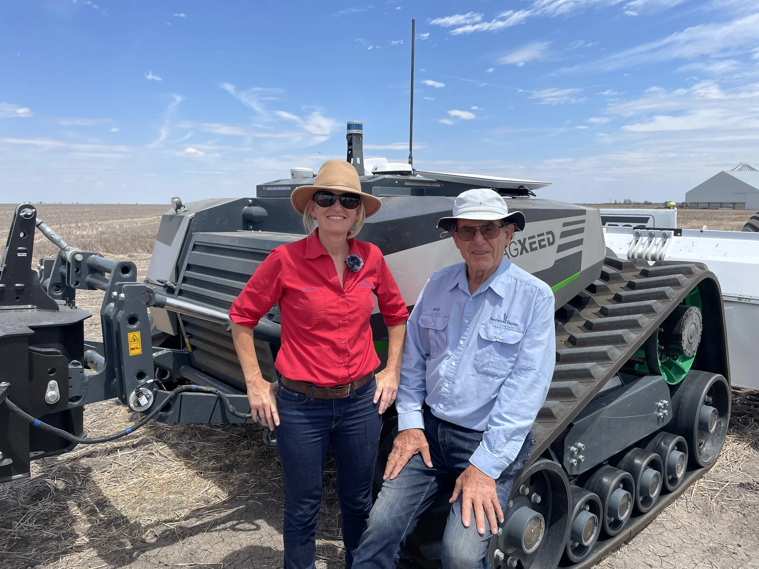 Two people standing by a large agricultural vehicle in a field, with one person wearing a wide-brimmed hat and sunglasses, and the other in a cap. The vehicle features caterpillar tracks and the word 'AGXeed' on its side, indicating advanced farming technology. The sky is clear with a few clouds.