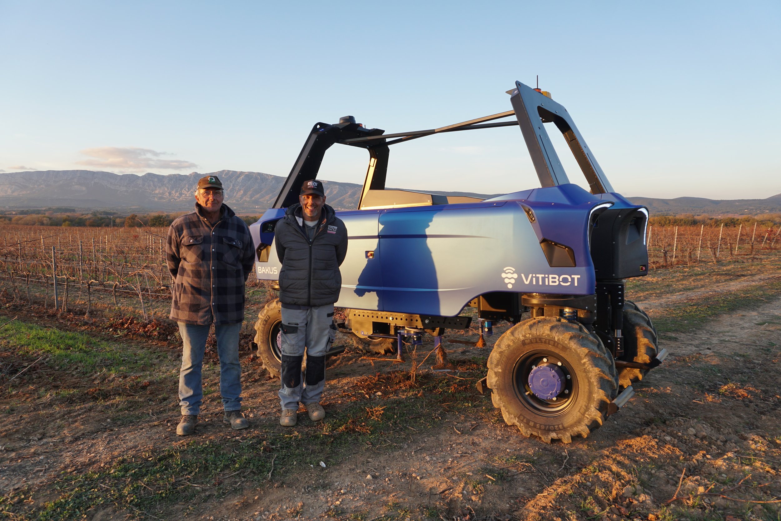 Two people standing next to a Vitibot robotic vineyard vehicle in a field with mountains in the background.