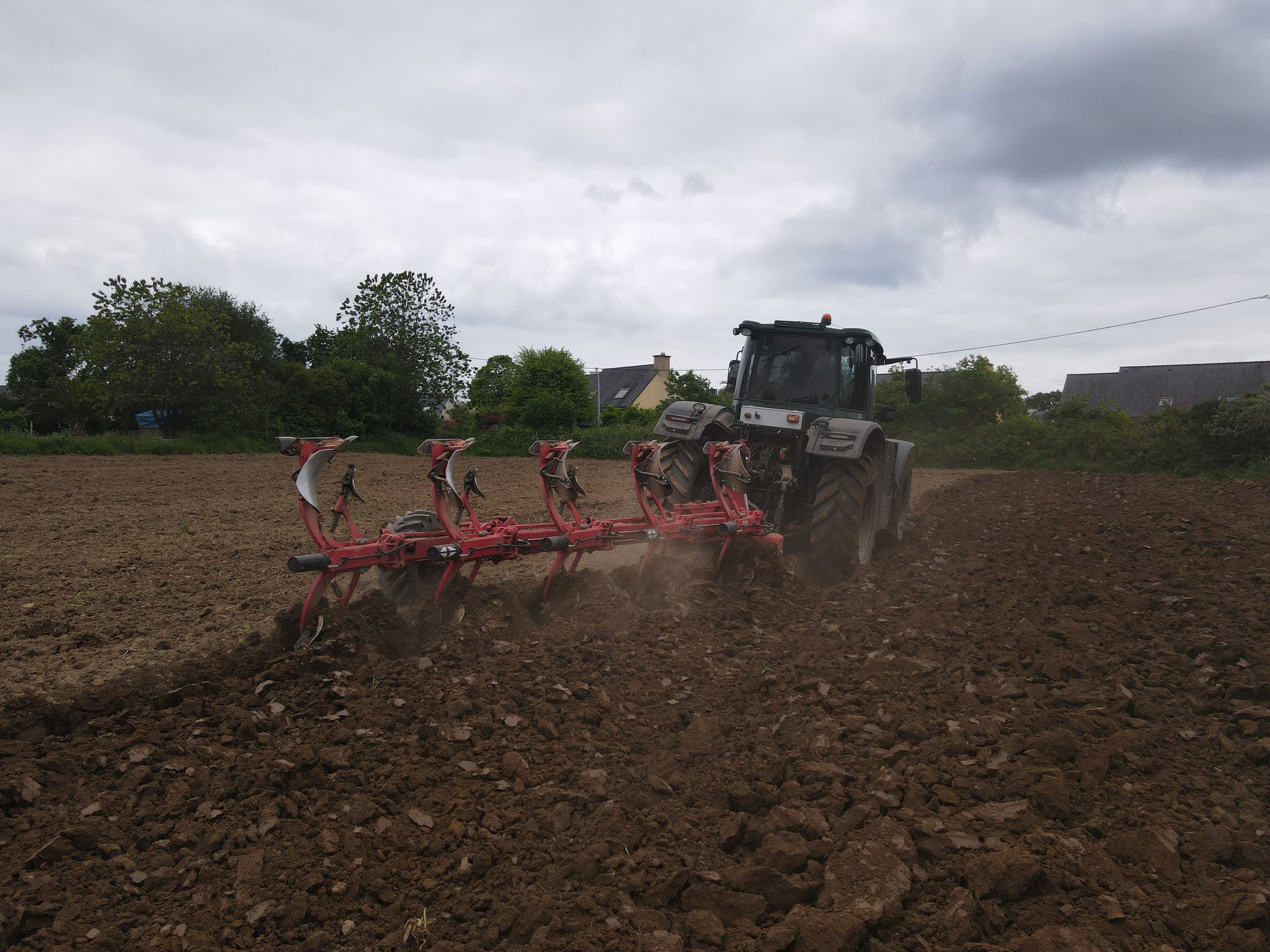 Tractor plowing a field with a red plow attachment, surrounded by trees and buildings under a cloudy sky.