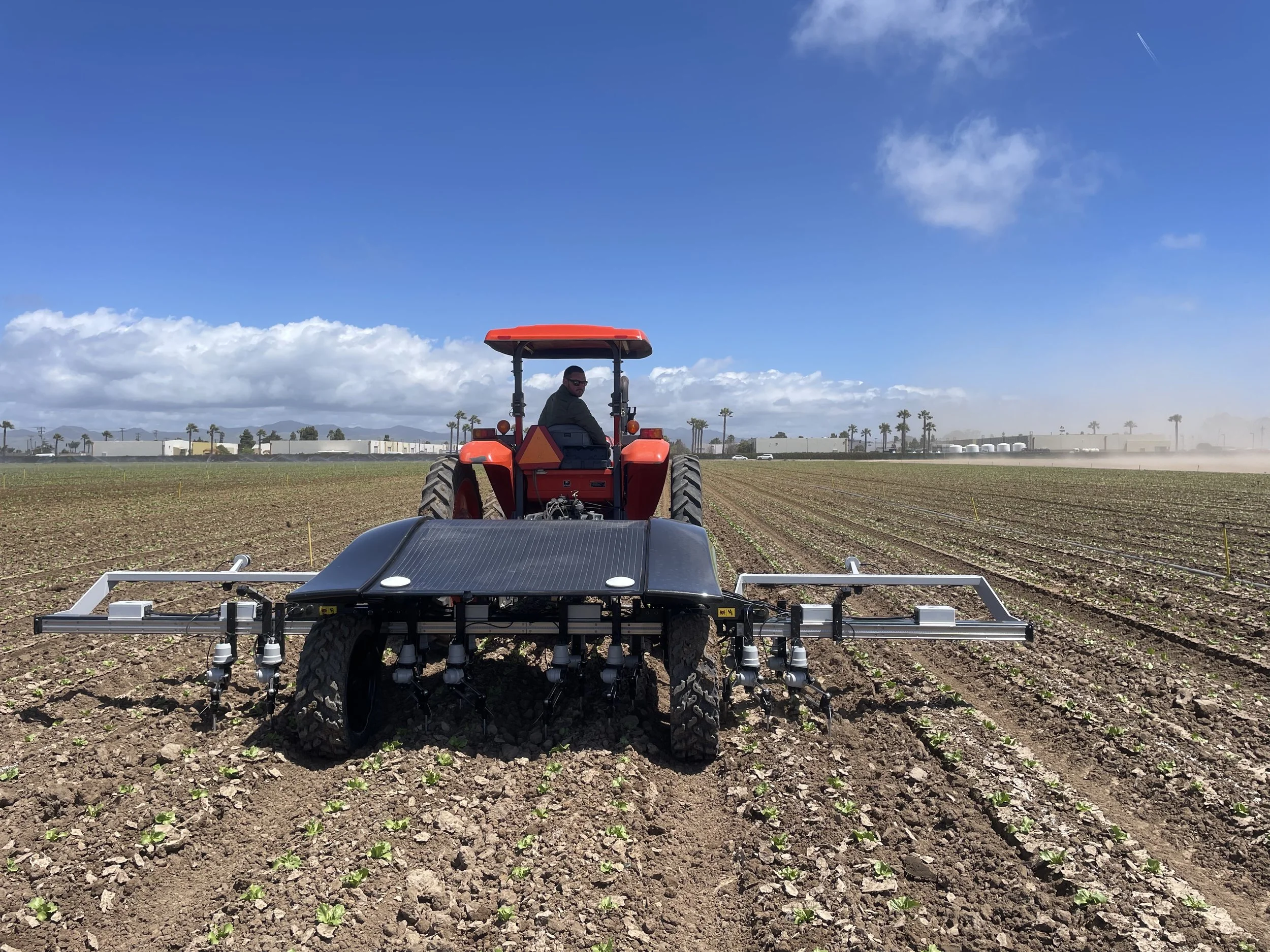 A farmer driving a red tractor equipped with a farming attachment in a newly planted field under a blue sky.