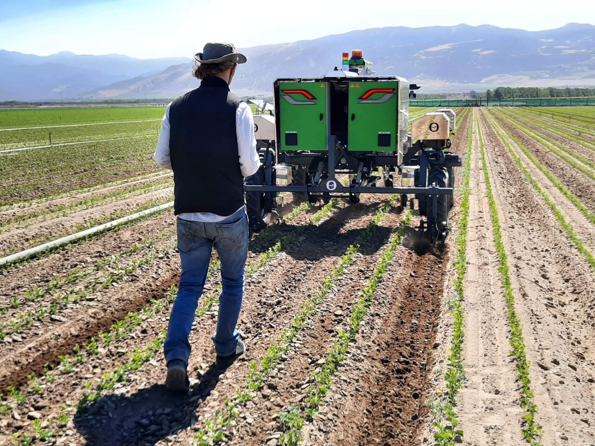 Person walking behind a green agricultural robot in a field, with mountains in the background.