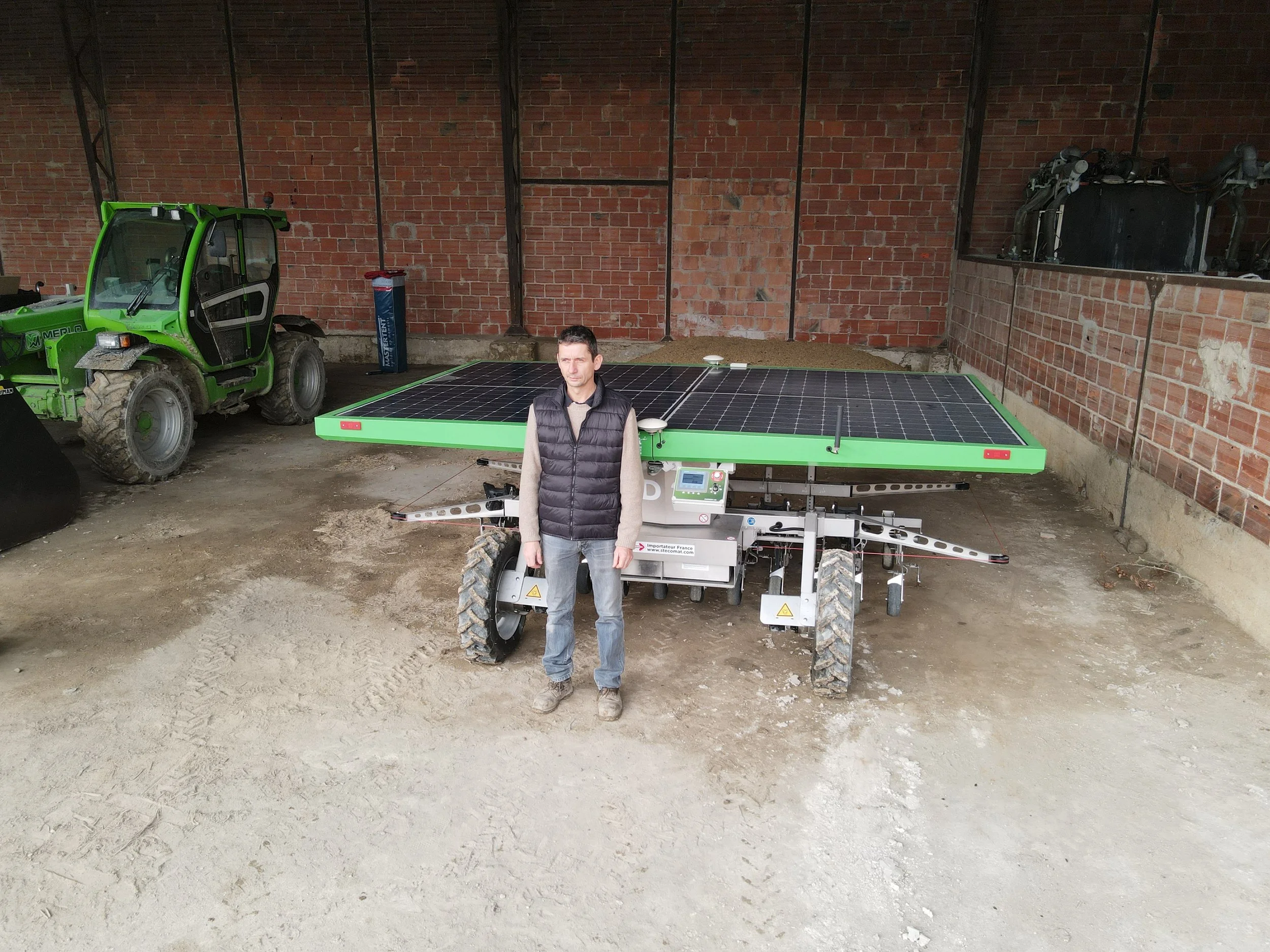 Man standing next to a large piece of equipment with solar panels, inside a brick-walled garage, with a green tractor in the background.