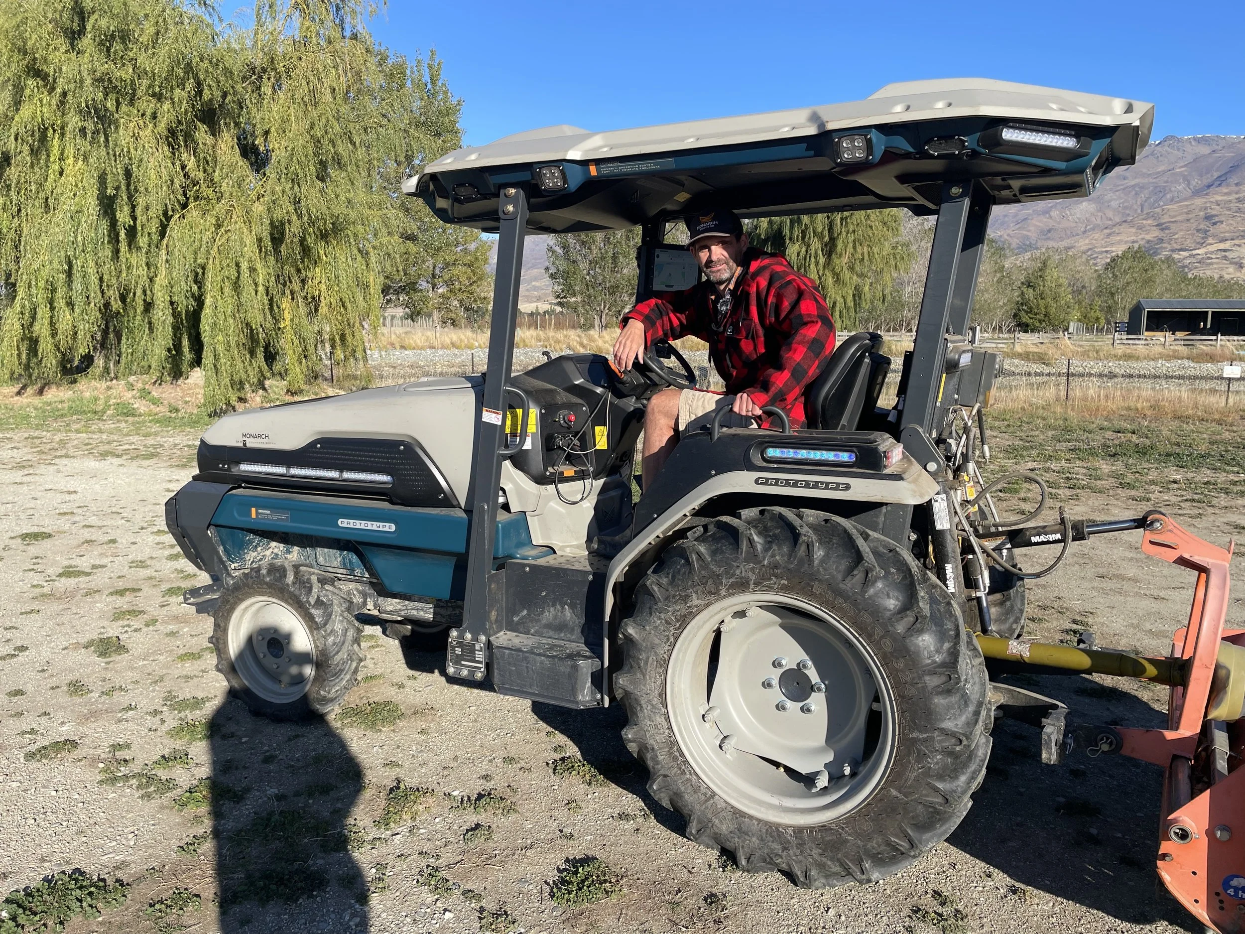 A person in a red and black plaid jacket sitting on a tractor in an outdoor setting with trees and mountains in the background.