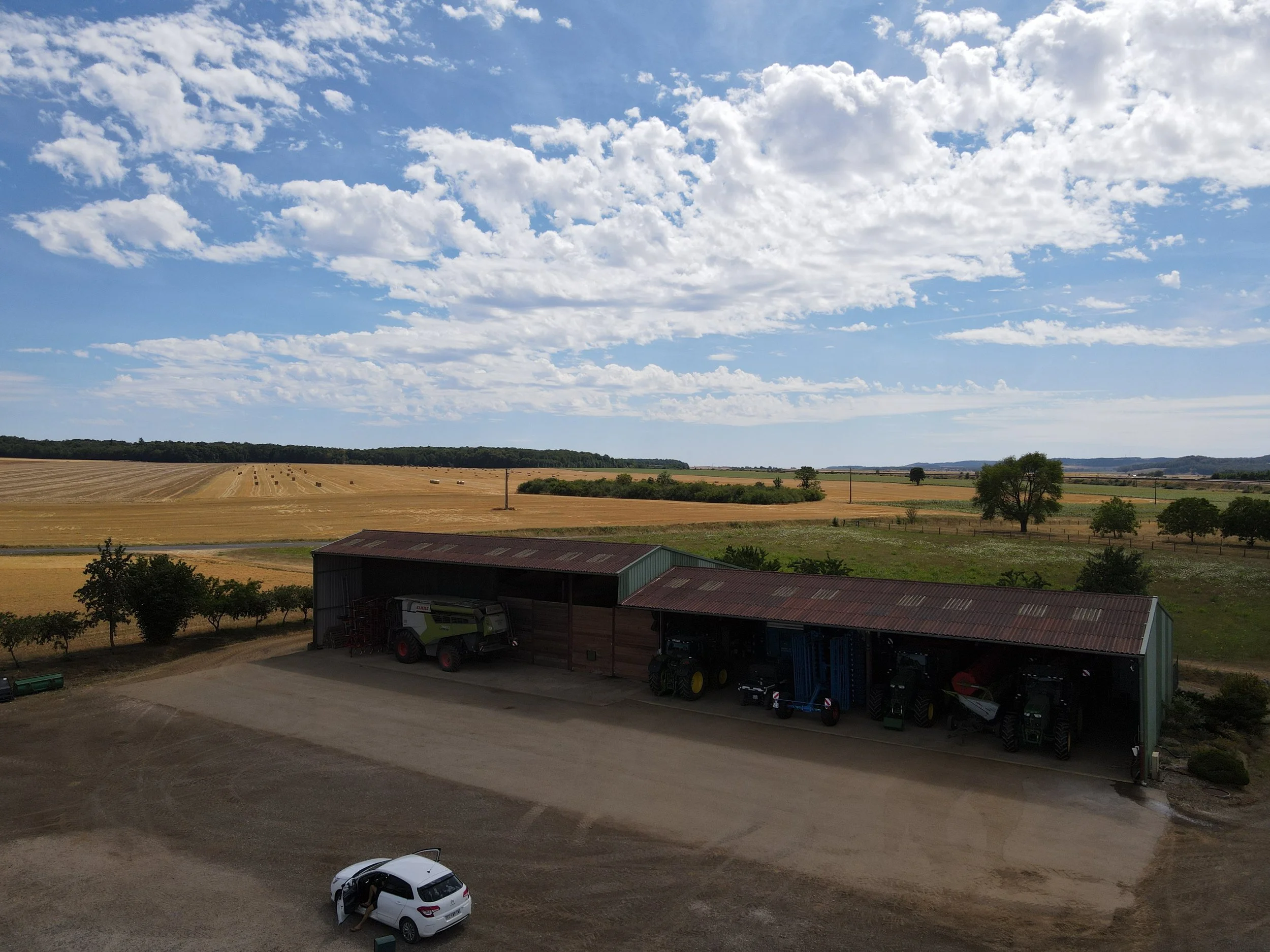 A rural landscape with a farm building housing agricultural machinery, a white car parked in front, and a vast expanse of golden fields under a partly cloudy sky.