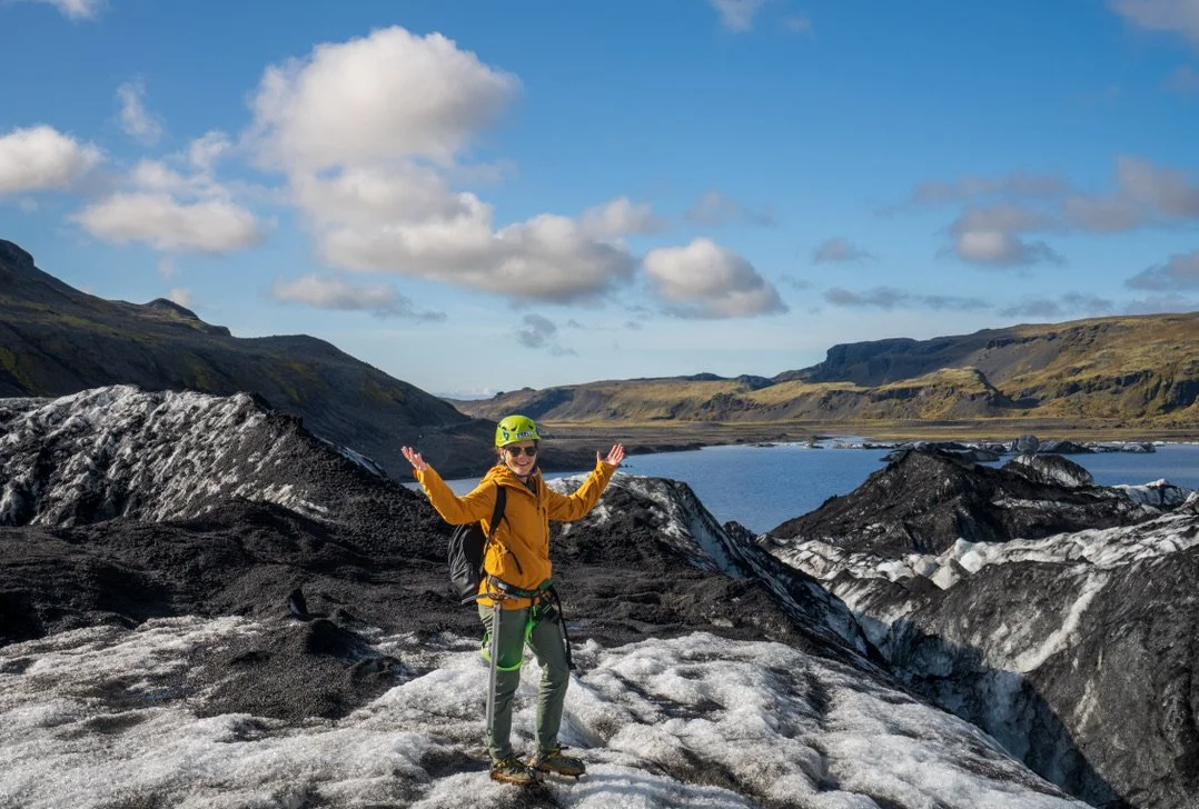 A woman in outdoor gear, including a yellow jacket and helmet, stands on a glacier with mountains, a lake, and a partly cloudy sky in the background.