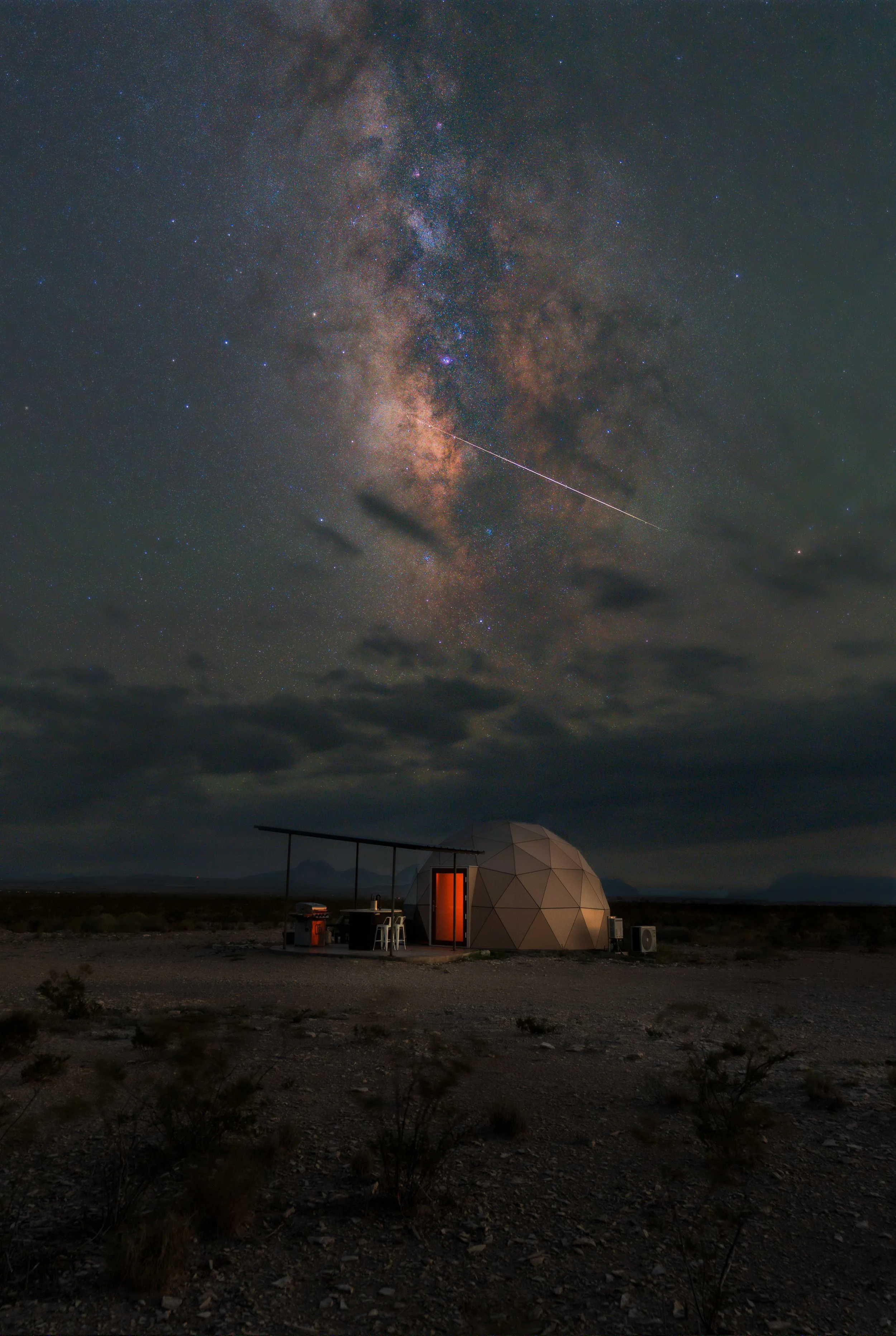 A geodesic dome-shaped tiny house with lit windows in a desert, under a night sky with star trails forming concentric circles around the North Star.