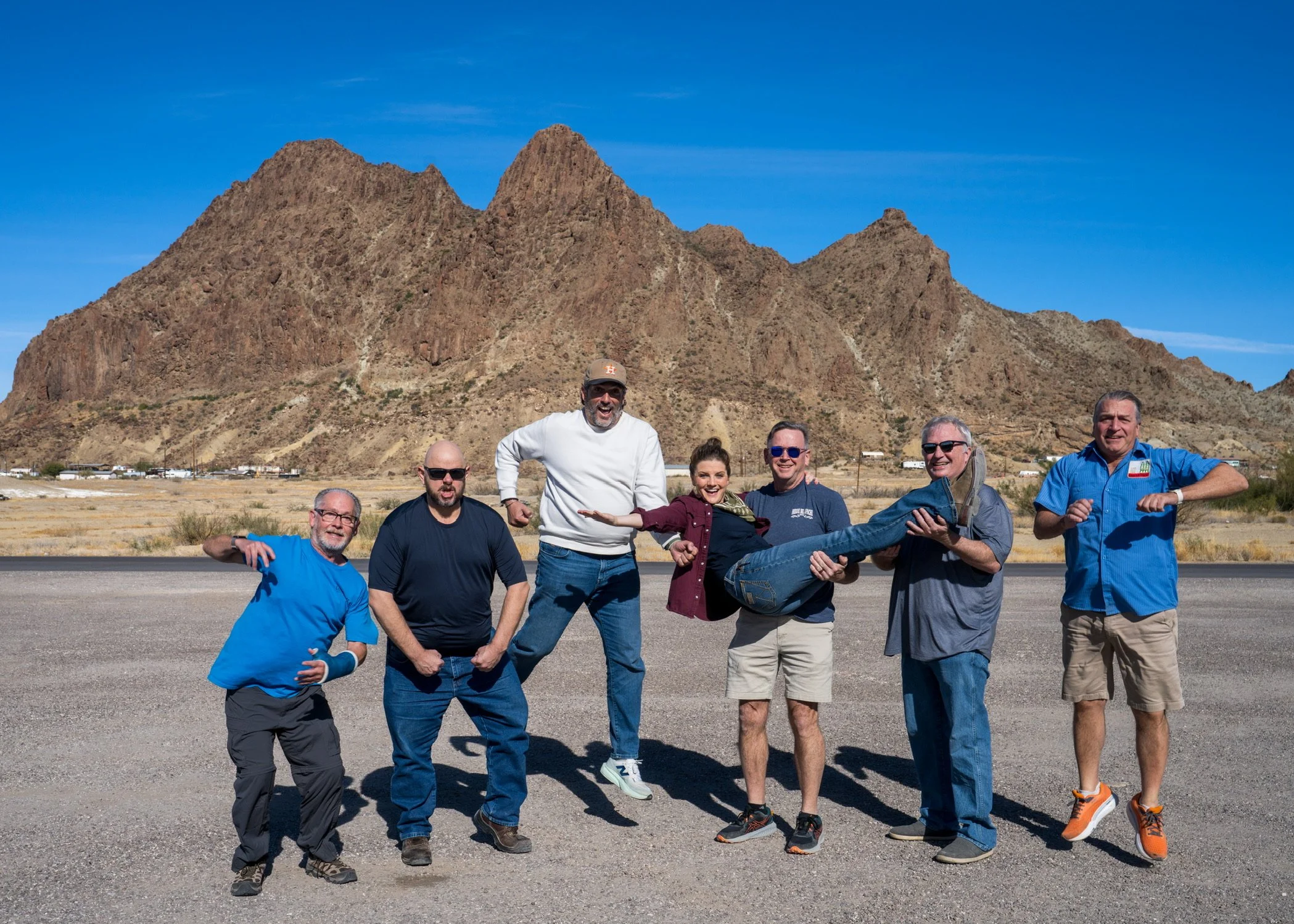 Group of seven people outdoors in a desert with mountains in the background, some holding a woman horizontally and smiling.