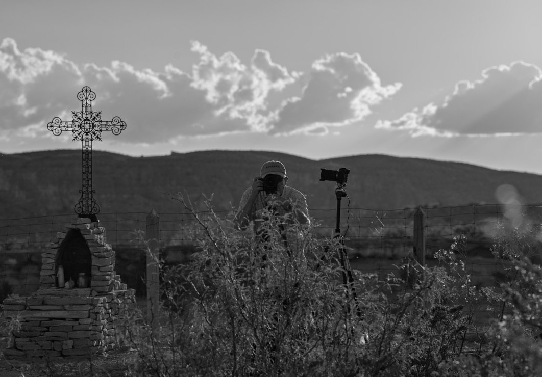 A person taking a photograph in a field with a decorative metal cross and a camera on a tripod, with mountains and cloudy sky in the background.