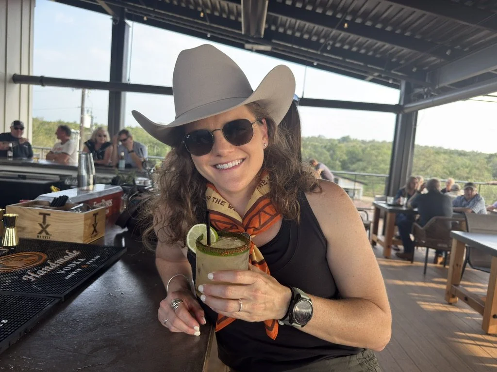 A woman wearing sunglasses, a cowboy hat, and a bandana around her neck, smiling and holding a margarita with a lime wedge in it at a bar or restaurant with other people seated in the background.