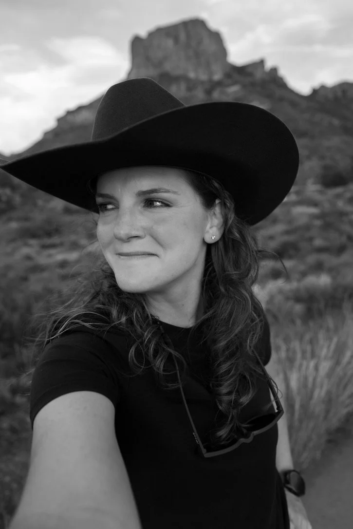 Black and white photo of a woman wearing a wide-brimmed hat, smiling slightly, with a rocky mountainous landscape in the background.