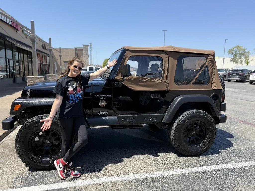 A woman poses next to a black Jeep Wrangler outdoors in a parking lot, smiling with her arms open, wearing sunglasses, a black t-shirt, leggings, and red and white sneakers.
