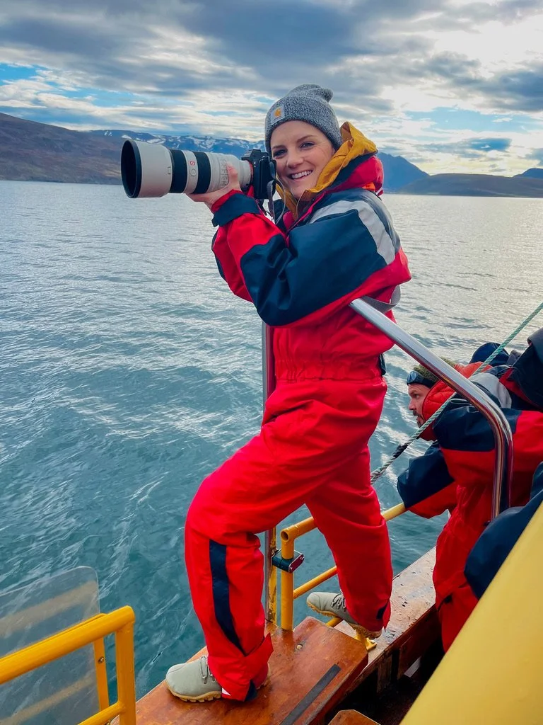 A young woman in red and blue waterproof gear, holding a camera with a large telephoto lens, onboard a boat on a lake surrounded by mountains, with a partly cloudy sky.