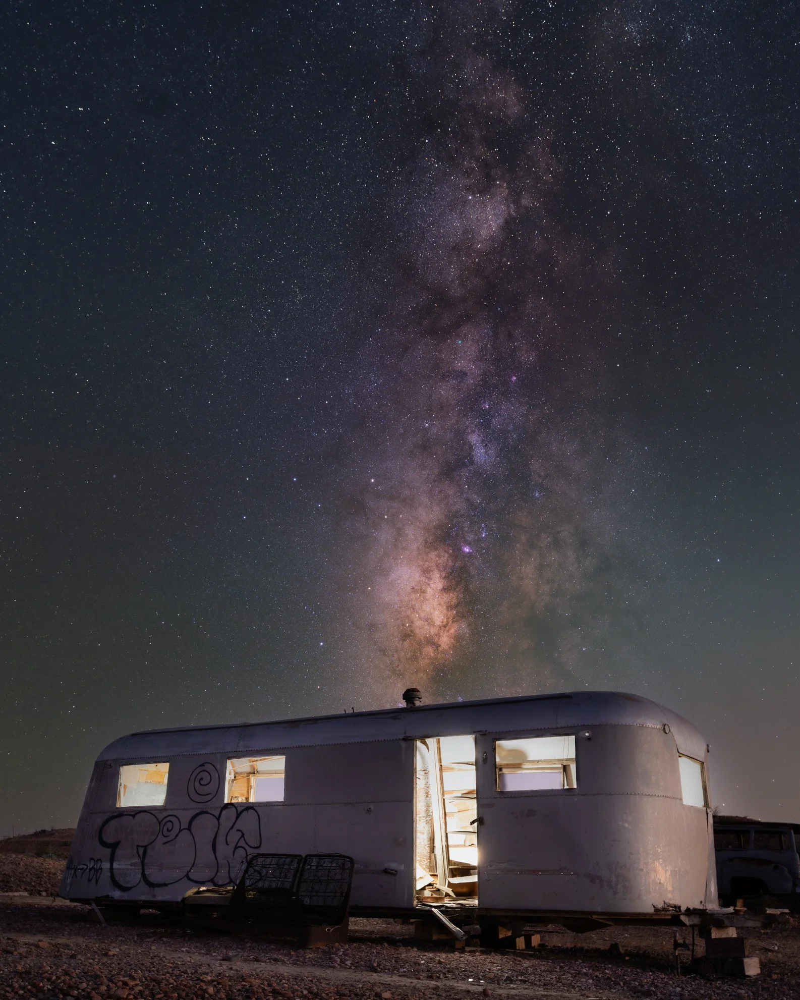 An old mobile home with graffiti on it, illuminated from inside, set against a night sky filled with stars and the Milky Way galaxy.