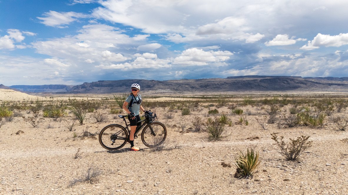 Person in helmet standing with a bike in a desert landscape with sparse desert plants, distant mountains, and a partly cloudy sky.