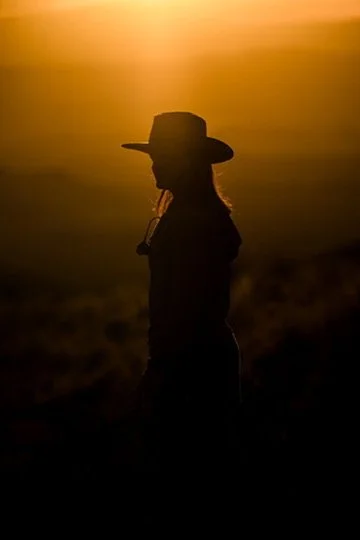 Silhouette of a woman wearing a wide-brimmed hat, standing outdoors during sunset or sunrise.
