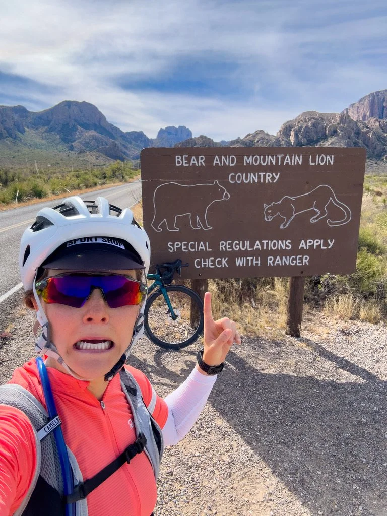 A woman in cycling gear with a helmet and sunglasses taking a selfie next to a sign that reads "Bear and Mountain Lion Country" in a desert landscape with mountains in the background.