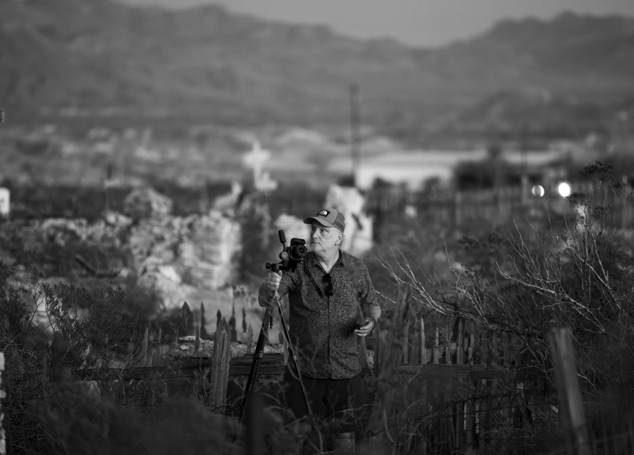 A man operating a camera on a tripod outdoors, with a rural landscape and mountains in the background, in black and white.