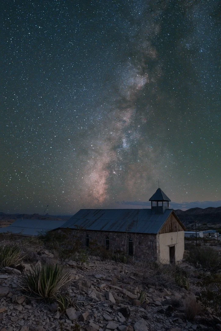 Nighttime scene of a stone chapel with a metal roof and small bell tower, set in a desert landscape with sparse vegetation, under a star-filled sky with the Milky Way galaxy visible.
