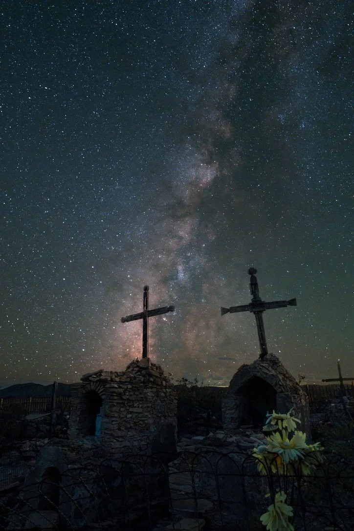 Night sky filled with stars and the Milky Way galaxy, with two cross-shaped structures and small stone buildings in foreground.