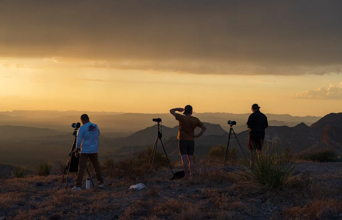 Three photographers with tripods capturing sunset over mountainous landscape.
