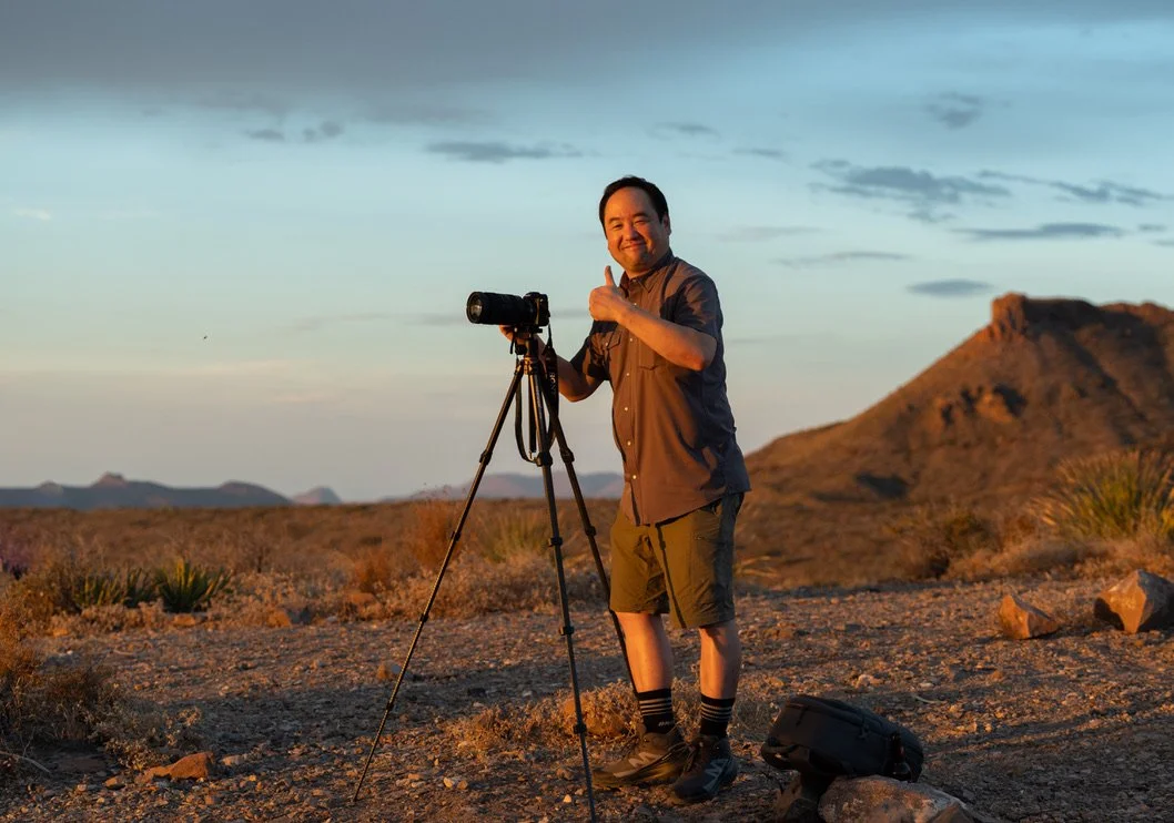 A man with a camera on a tripod giving a thumbs up in a desert landscape during sunset.