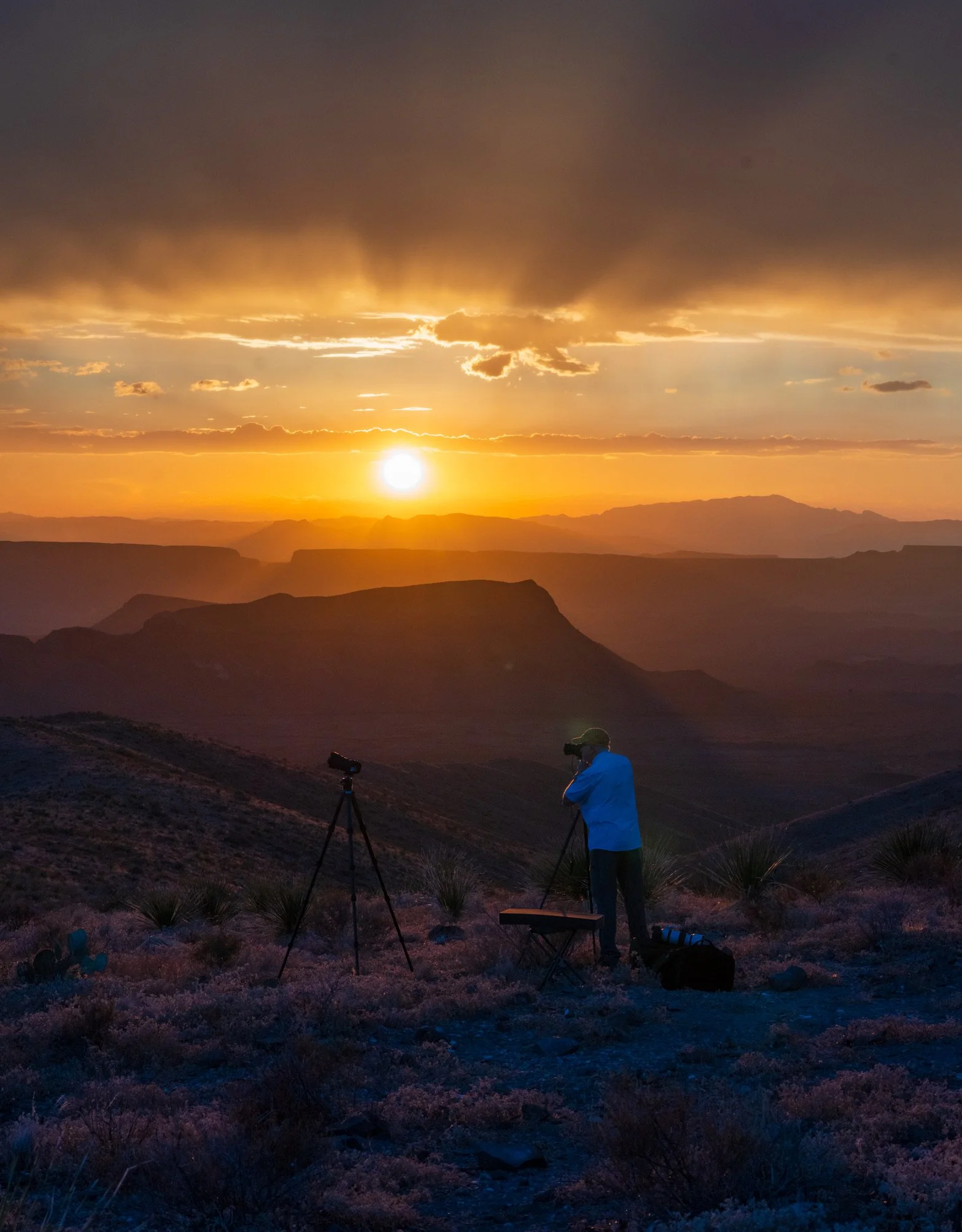 A person taking photographs of a sunset in a desert landscape with mountains and rocky terrain.