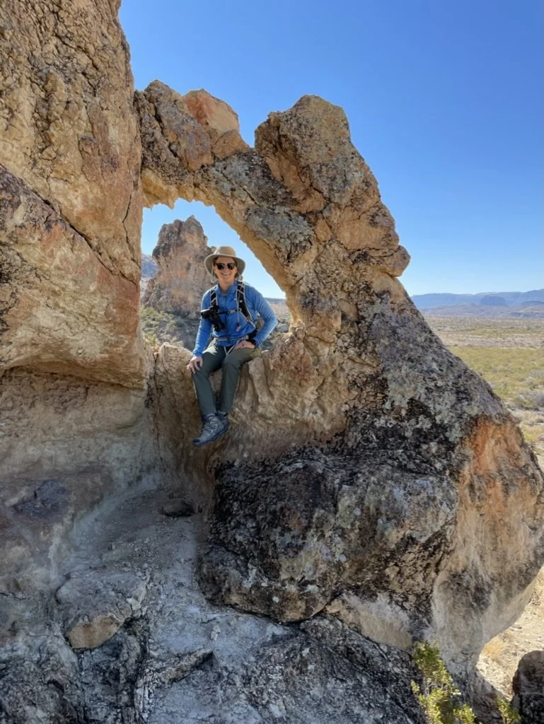 A woman with a hat, sunglasses, and a blue shirt sitting on a large rock formation with a natural arch, in a desert landscape with mountains in the background under clear blue sky.