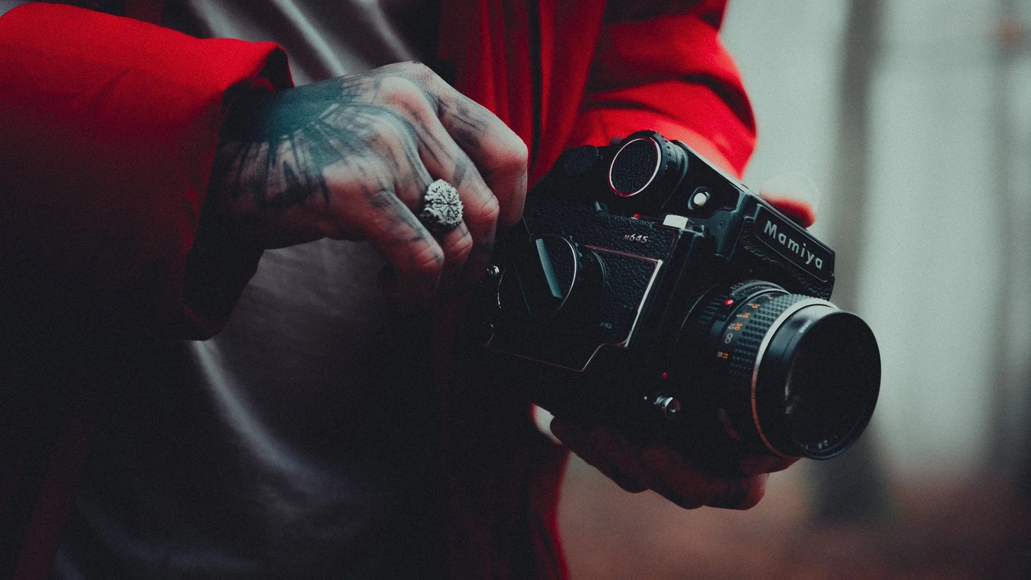 Person holding a vintage film camera, wearing a red jacket and a silver ring with a star design, tattoos visible on hand.