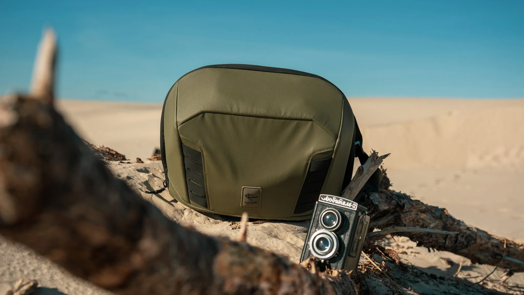 A green backpack and a vintage camera with a sticker that has Russian text sit on a sandy beach near driftwood, with a clear blue sky in the background.