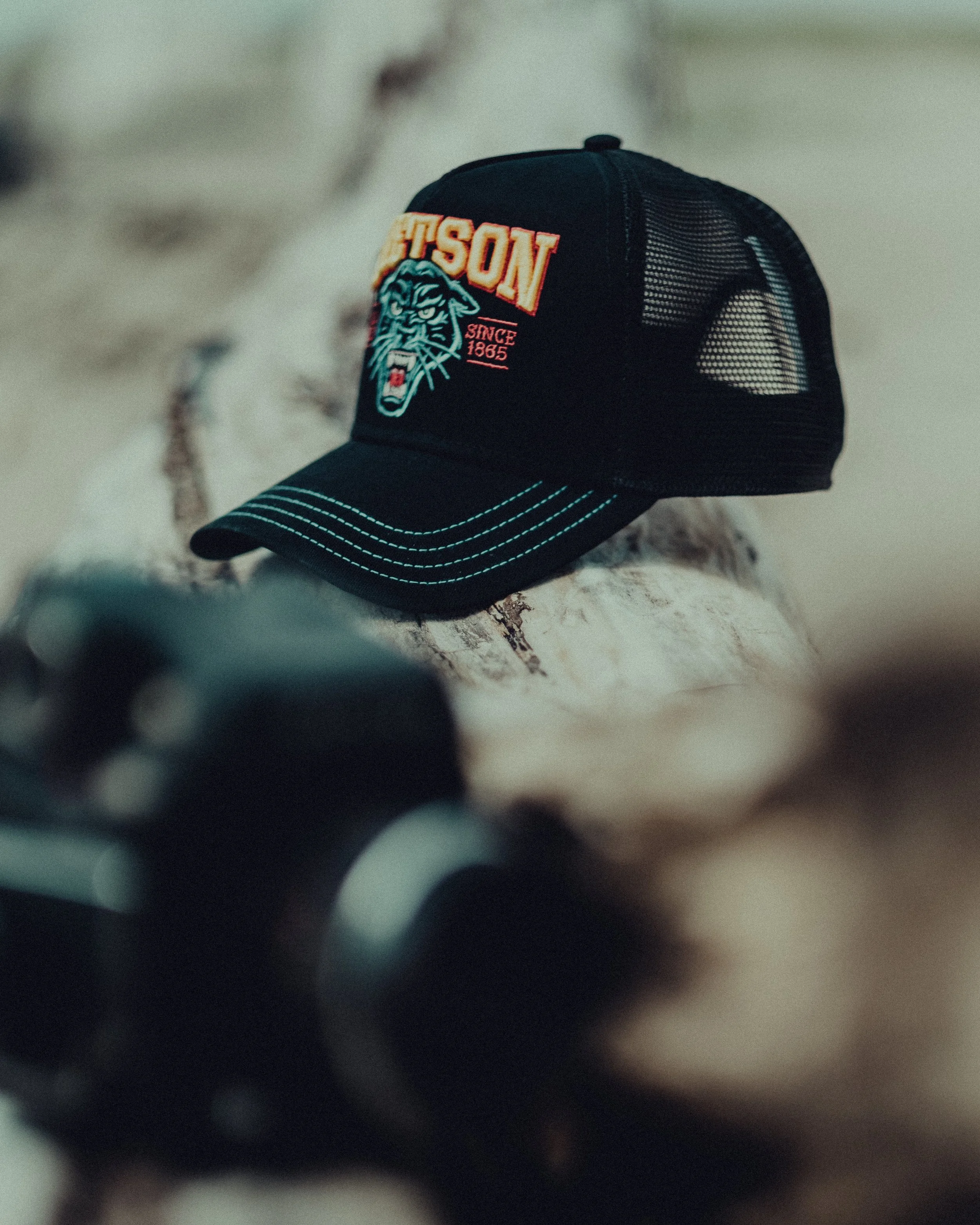A black trucker cap with embroidered logo and text, resting on a rock, with a blurred object in the foreground.