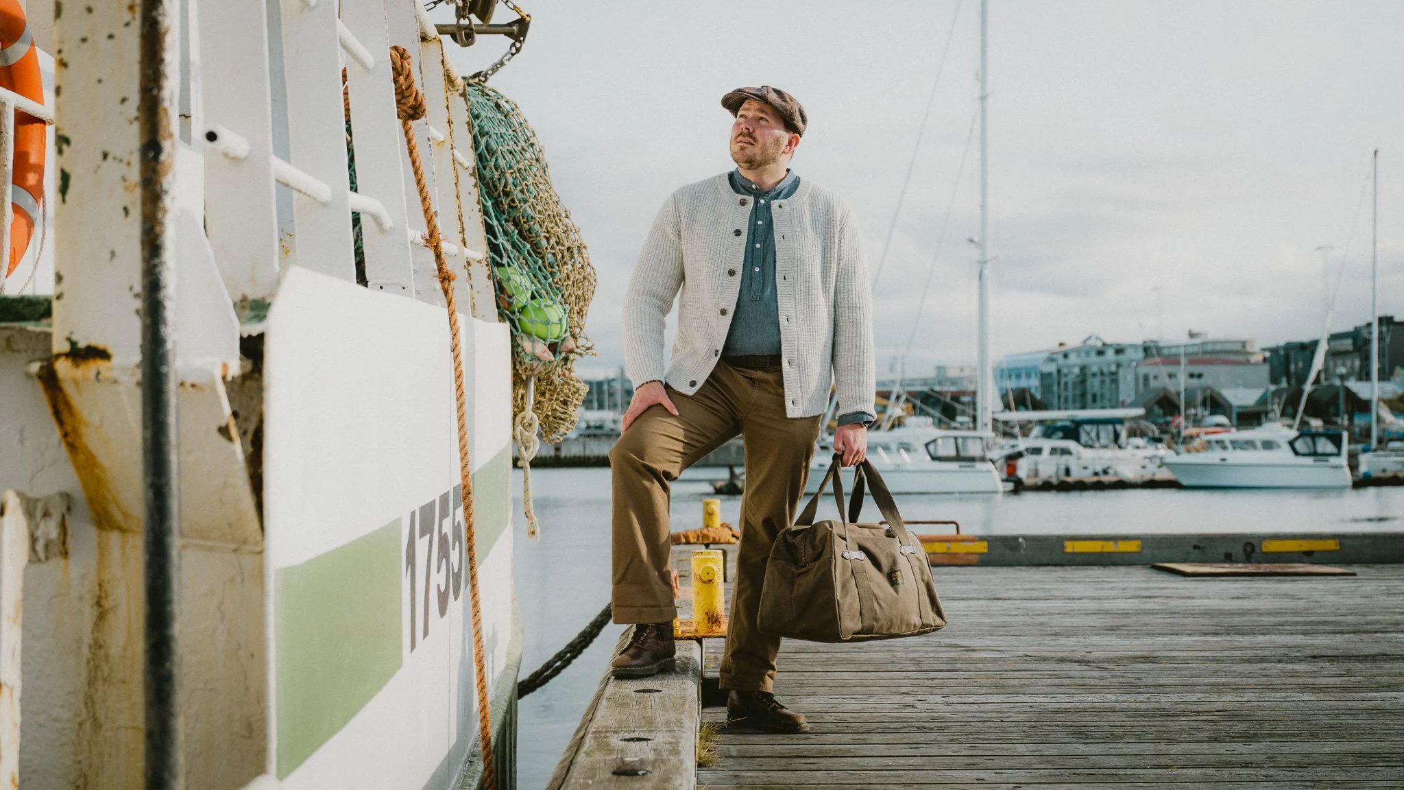 A man with a beard and flat cap stands on a dock at the marina, holding a duffel bag, with boats and buildings in the background.