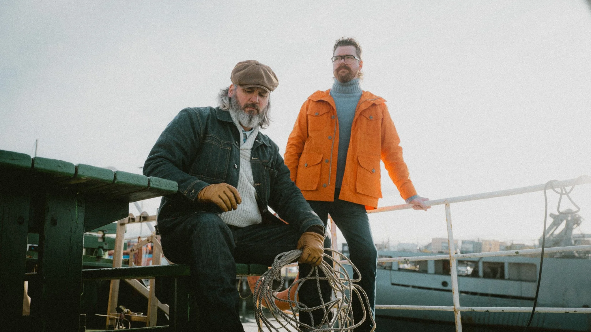 Two men near a boat dock, one sitting with a rope and the other standing with a railing, in a harbor or marina setting with boats and buildings in the background.
