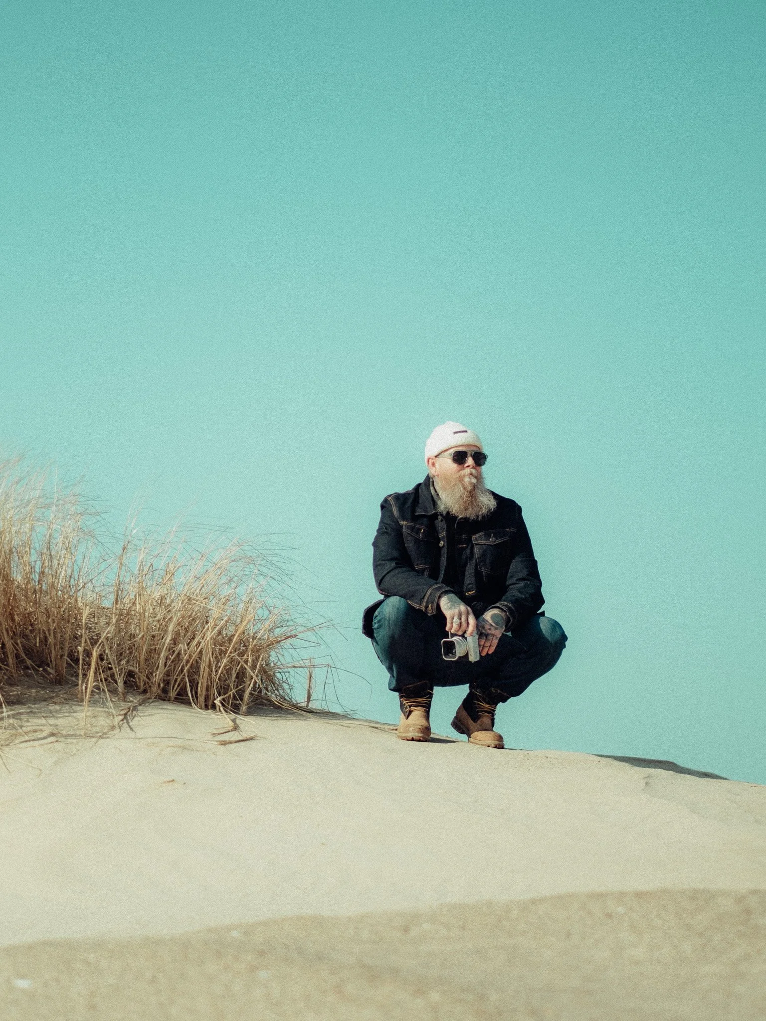 A man with a beard wearing sunglasses, a white beanie, a denim jacket, and brown boots squatting on a sandy beach, holding a camera, with dried grass and a clear blue sky in the background.