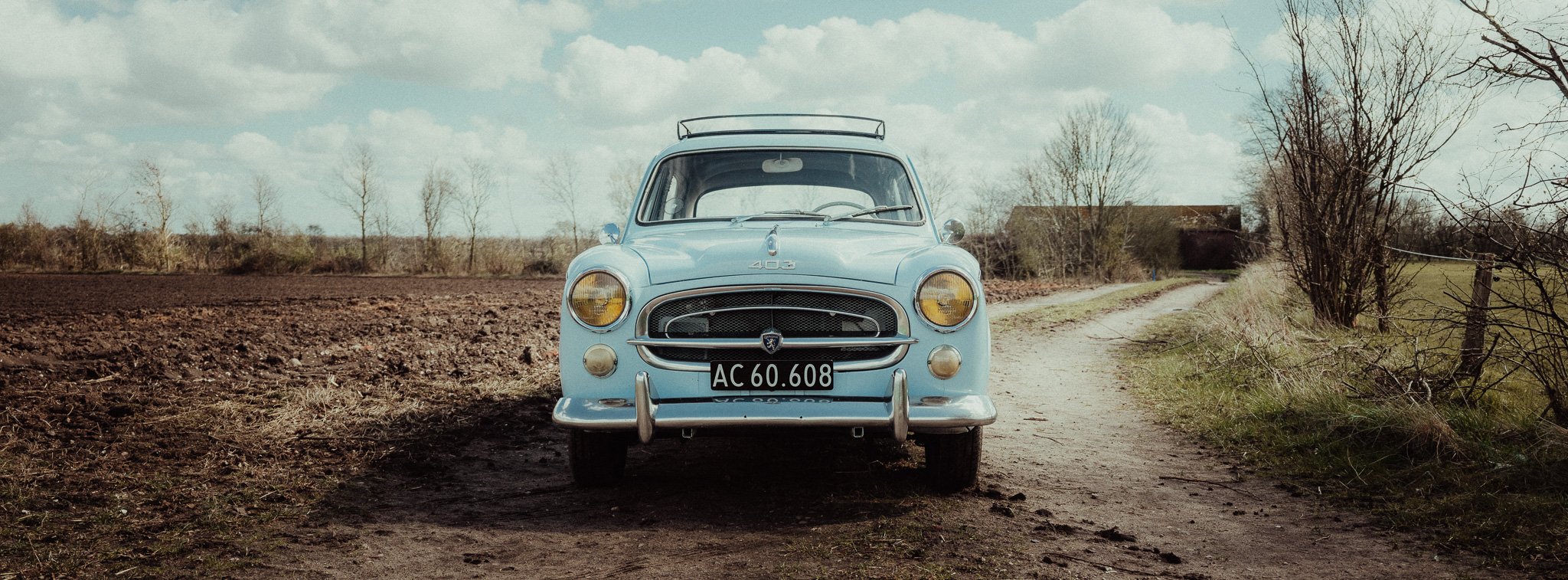Light blue vintage car parked on a rural dirt road with fields and trees in the background under a cloudy sky.