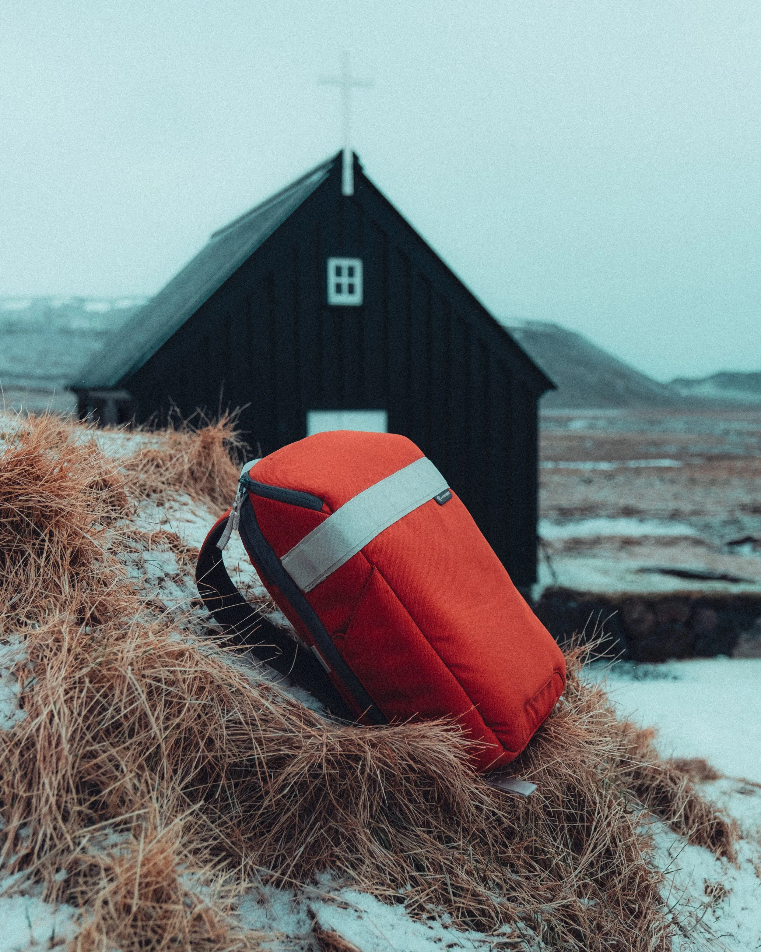 Red backpack resting on dry grass in front of a small black church with a white cross on top in a cold, overcast landscape.