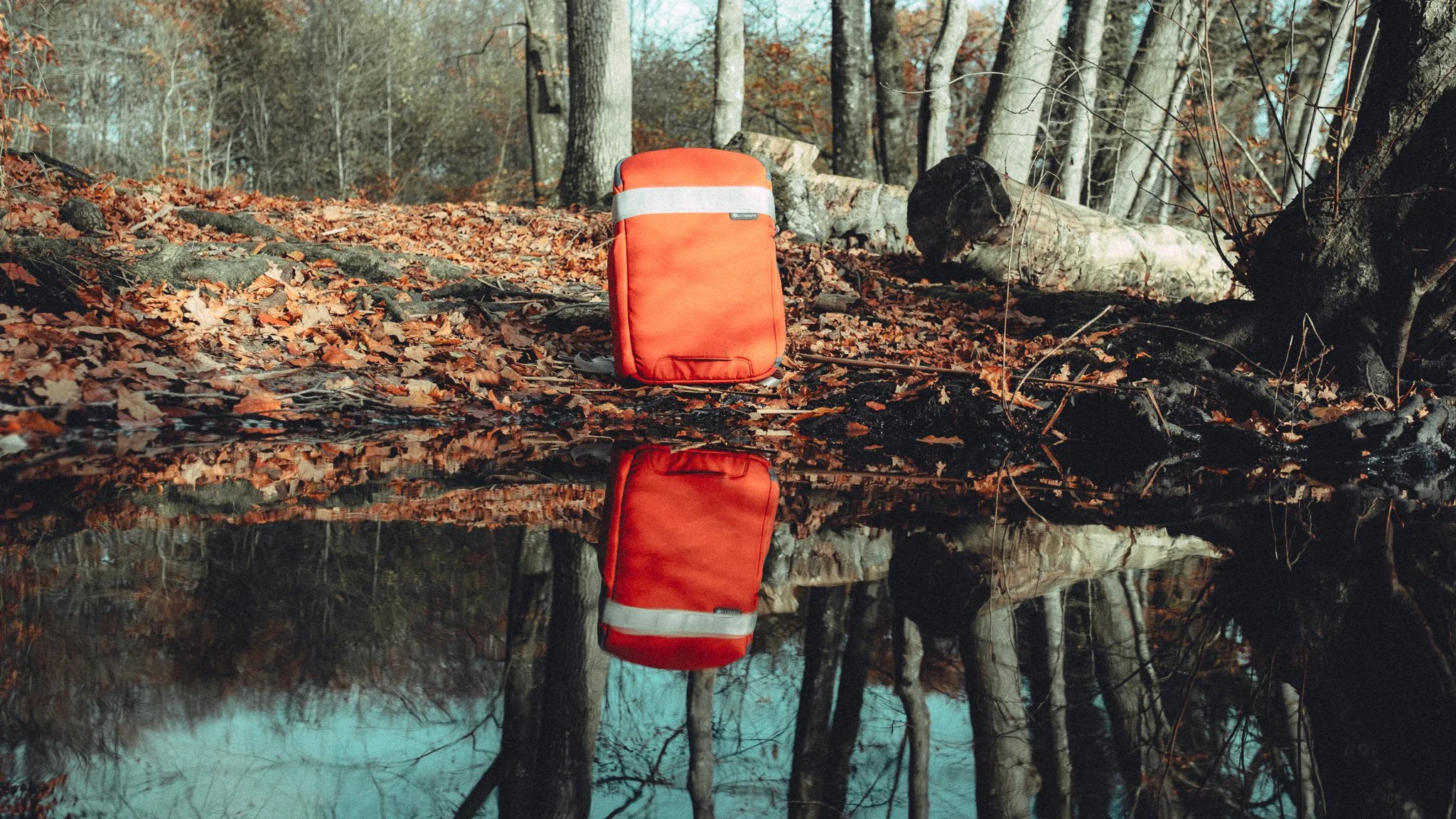 An orange backpack with a reflective stripe on a forest floor covered with fallen leaves, next to a small creek, with trees in the background, and a reflection of the backpack and trees in the water.