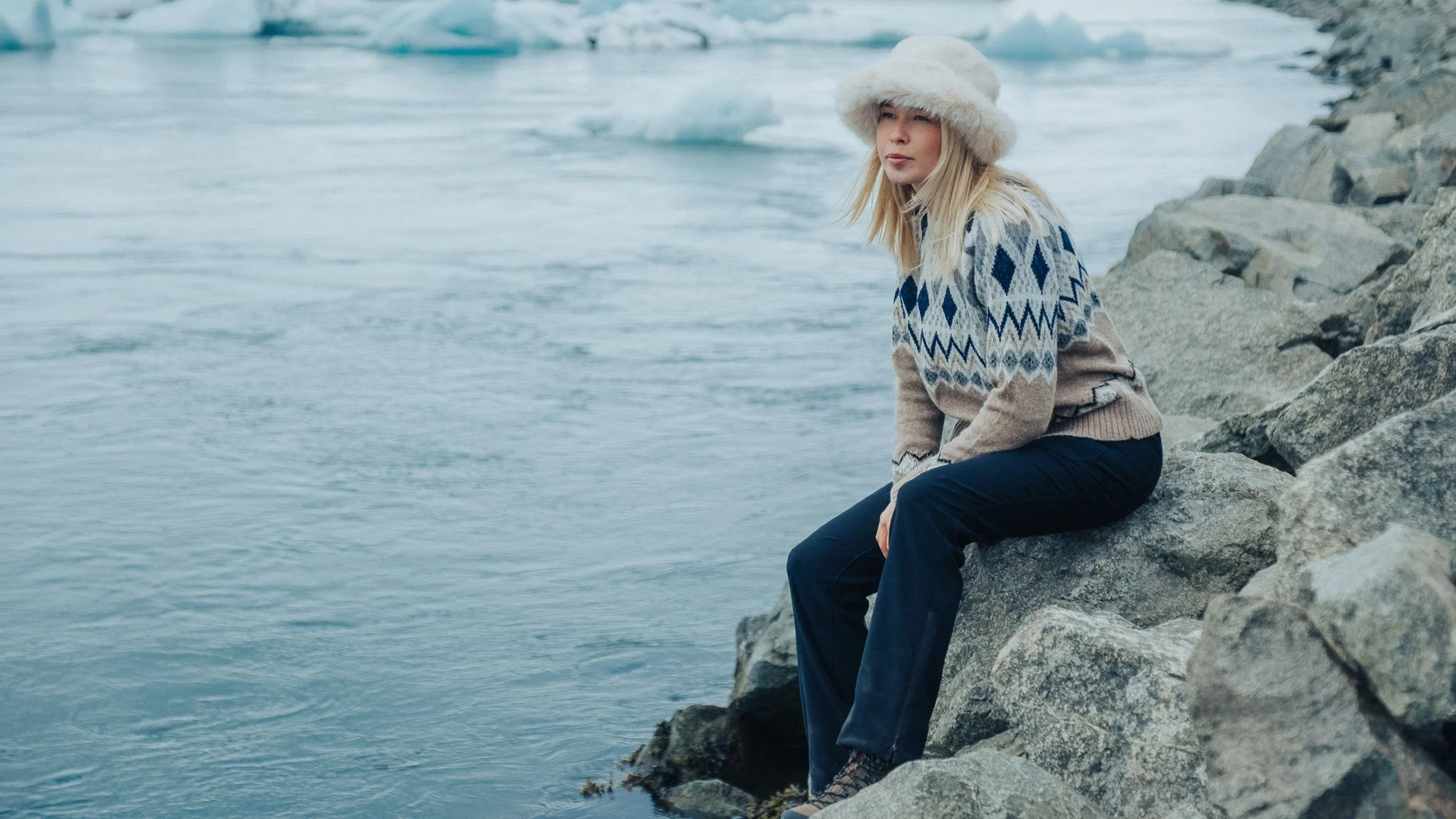 A woman wearing a fur hat and patterned sweater sitting on rocks by cold water with icebergs in the background.