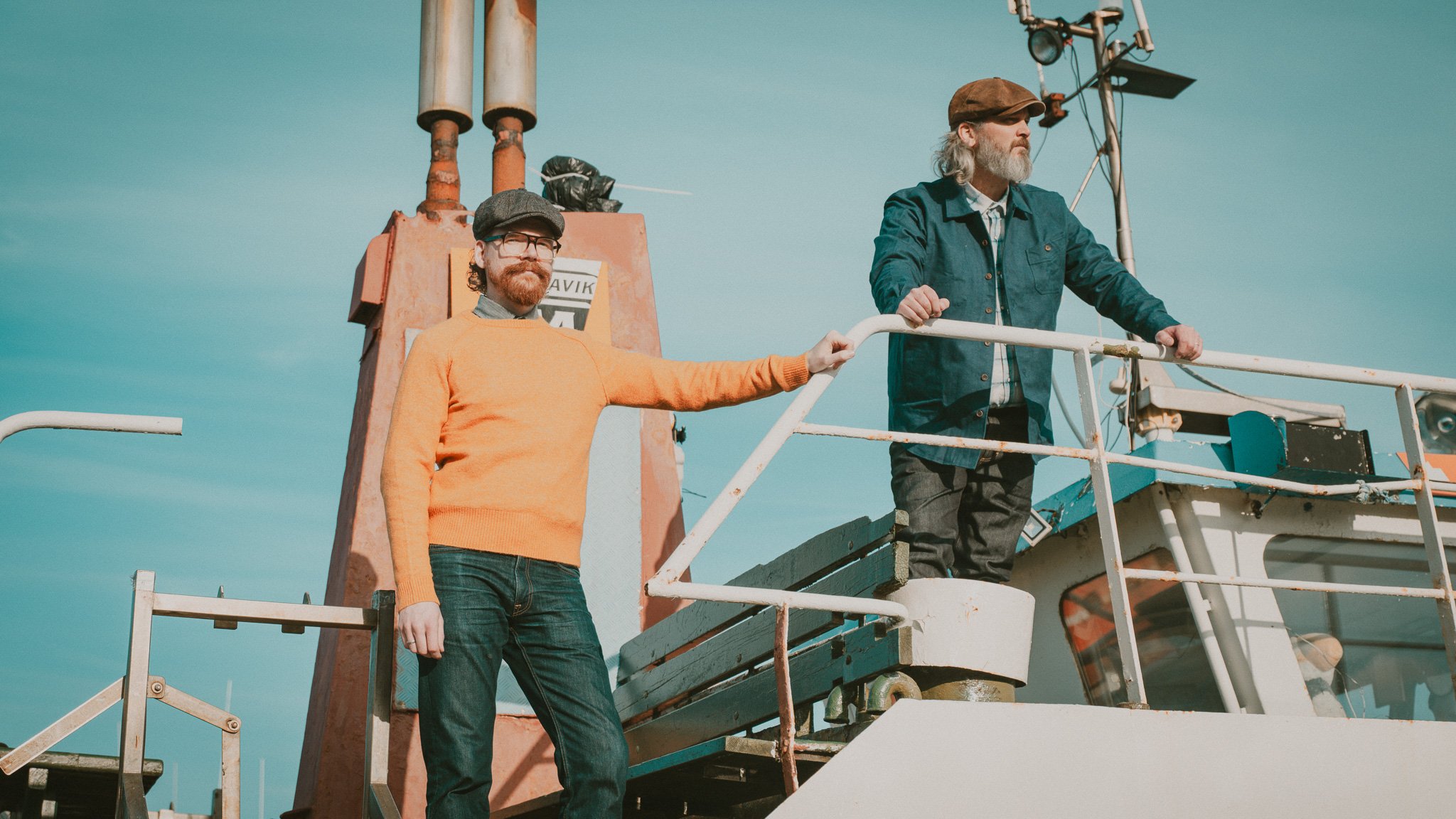 Two men on a boat, one in an orange sweater and glasses, the other in a blue jacket and hat, standing on the boat's deck with a clear blue sky in the background.