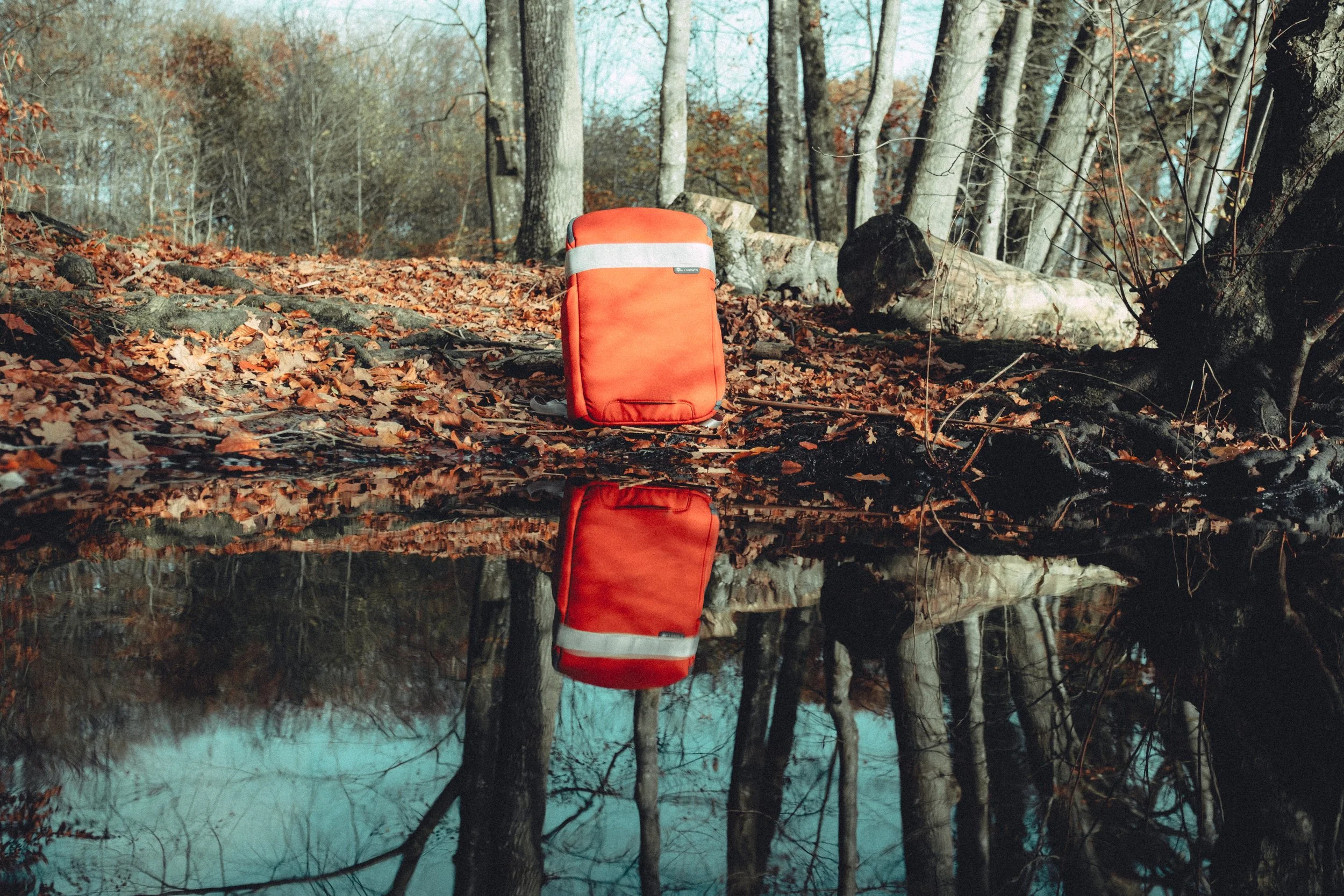 A red backpack with a reflective stripe sits on a leaf-covered forest ground near a small pond, with the pond's reflection showing the backpack and trees.