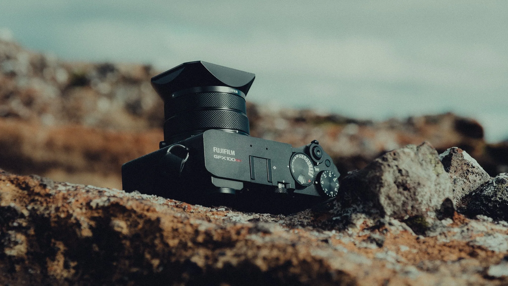A black digital camera with a lens, resting on rocky terrain outdoors.