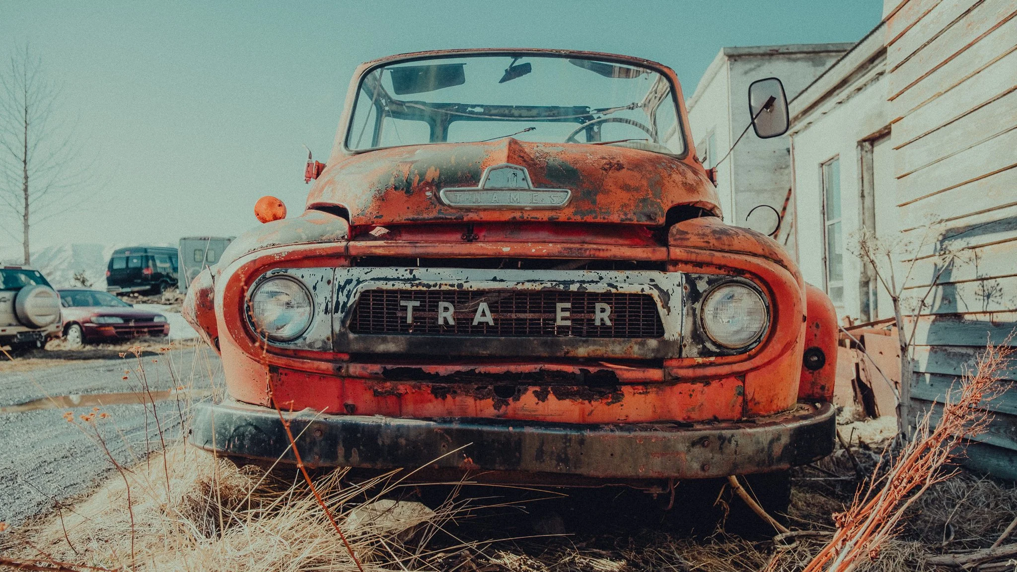 An old, rusty red truck with 'TRAYER' on the front grille, parked outdoors near a white wooden building with weathered siding, surrounded by other classic cars and dry grass.