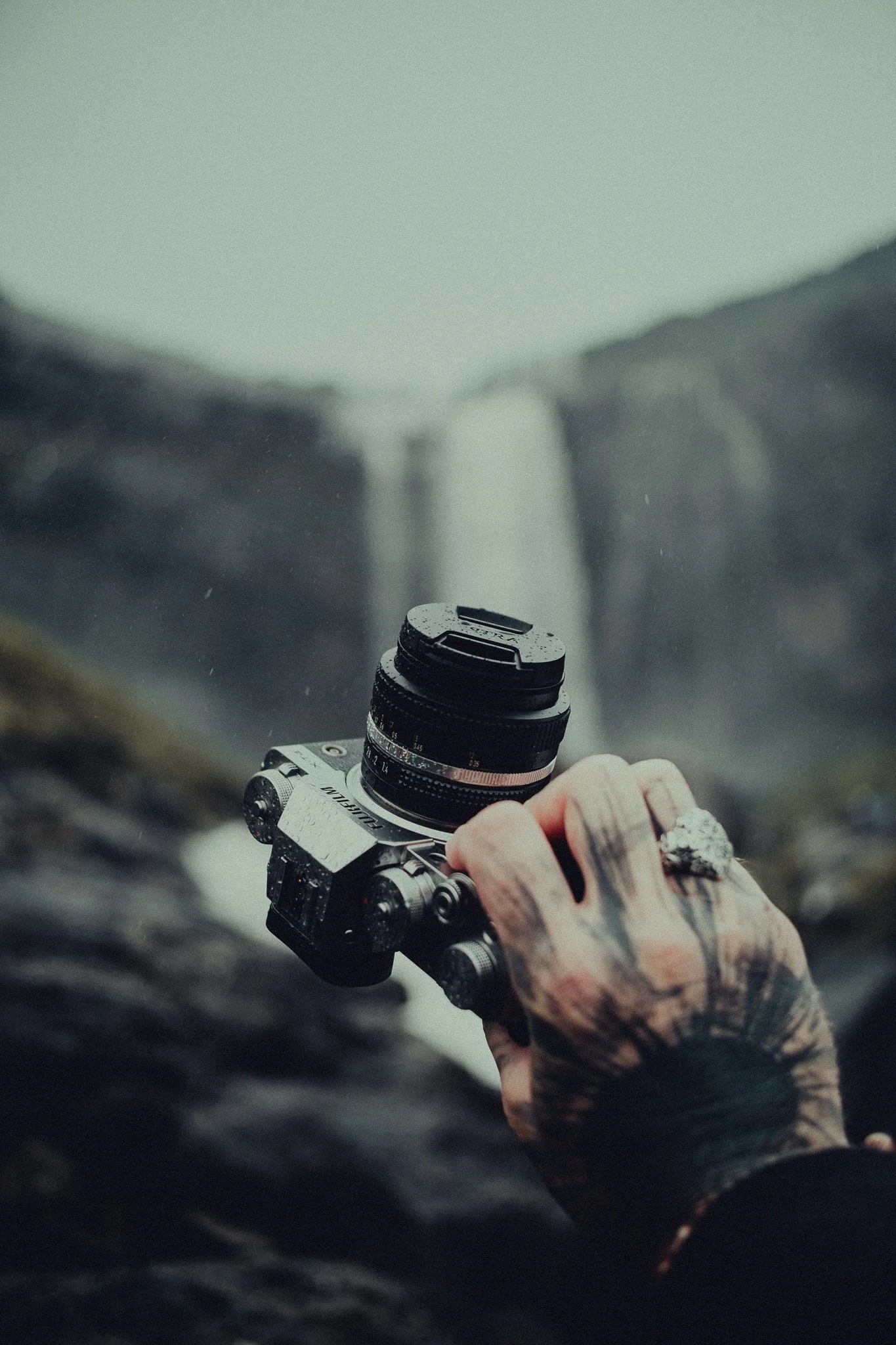 A tattooed hand holding a camera in a natural outdoor setting with a waterfall in the background.