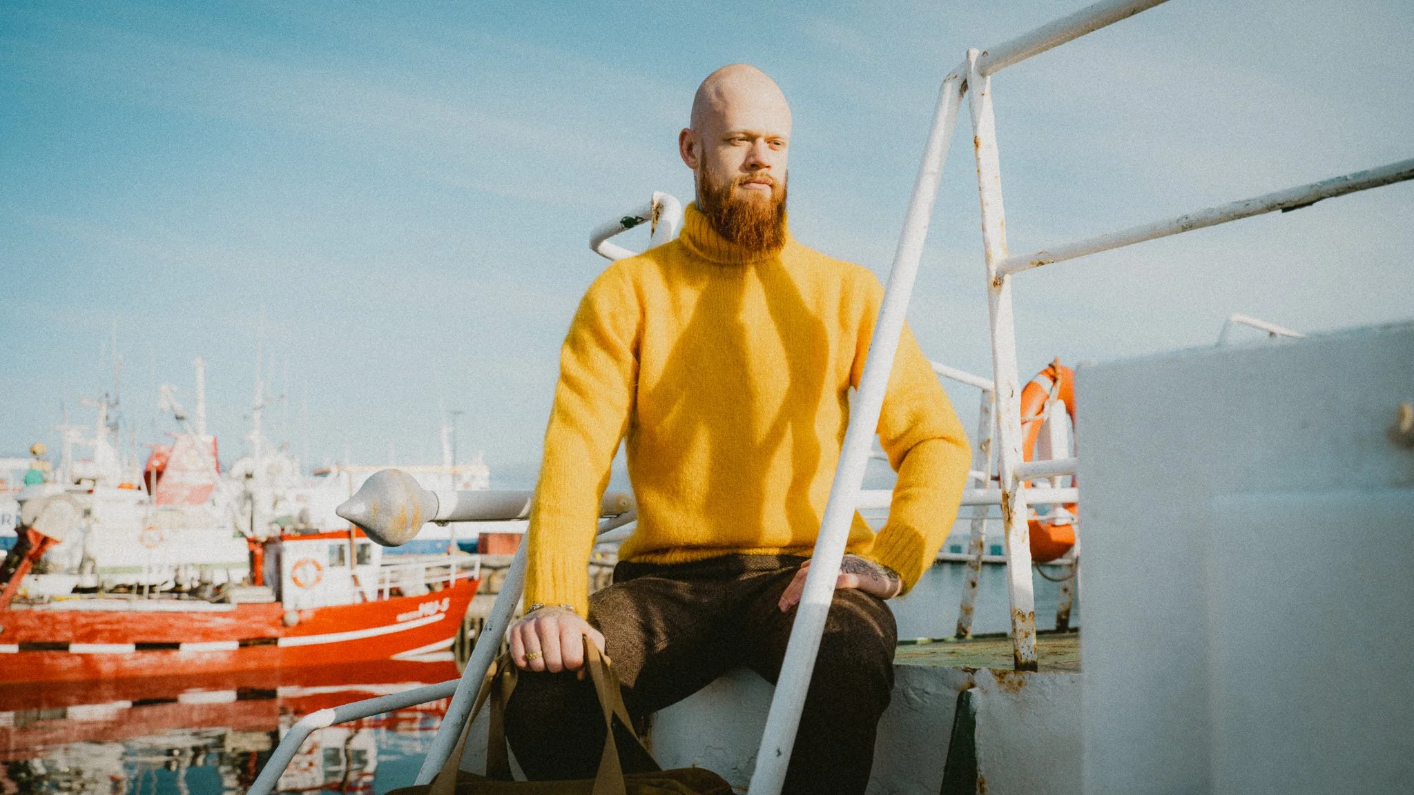 A man with a bald head and red beard wearing a yellow turtleneck sweater, sitting on a boat at a dock with several boats in the background under a blue sky.