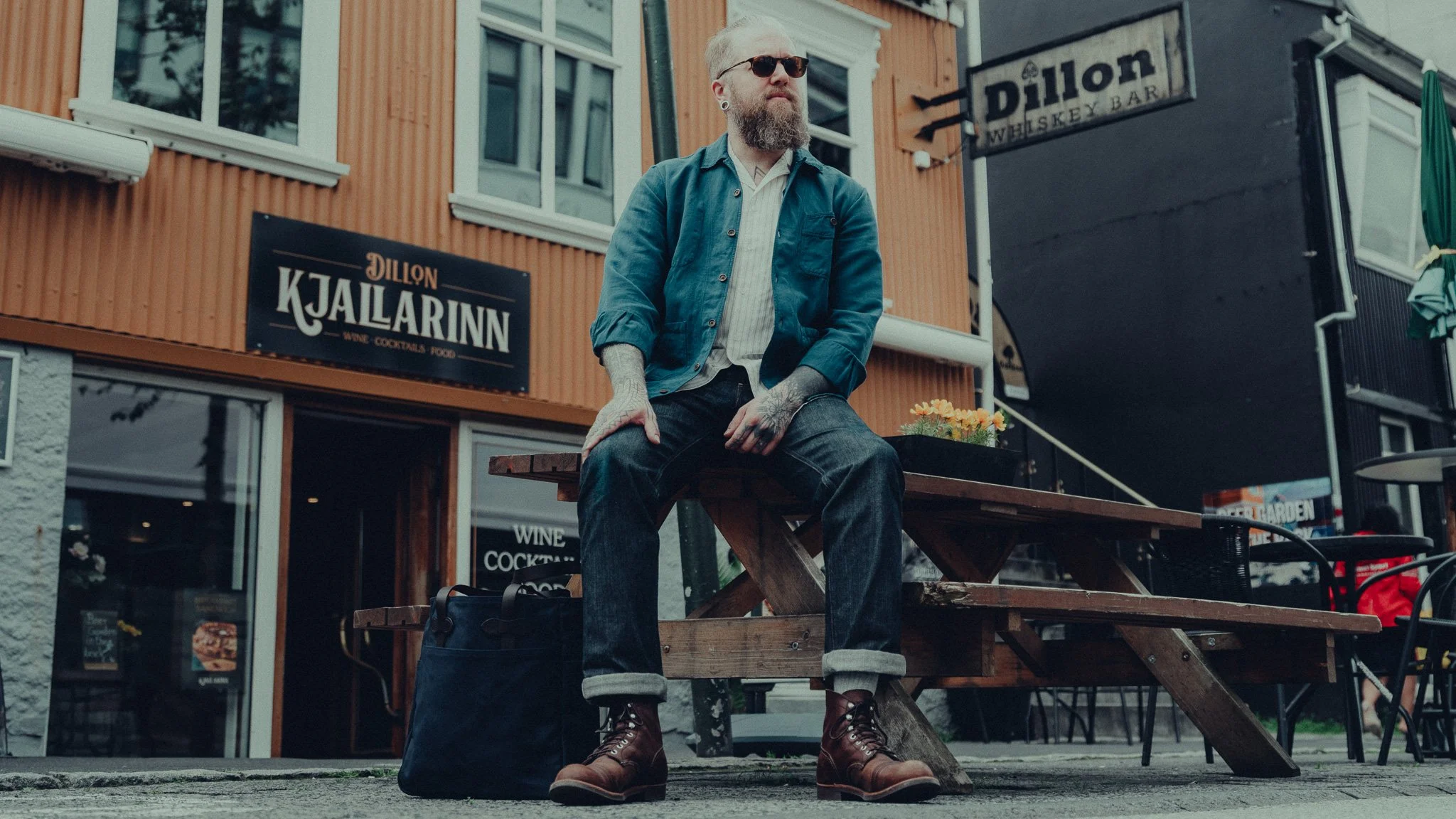 A man with a beard, sunglasses, and tattoos is sitting on a wooden bench outside a building with signs for Dillon Kjararinn and Dillon Whiskey Bar. He is wearing a blue jacket, white shirt, dark jeans, and brown boots.