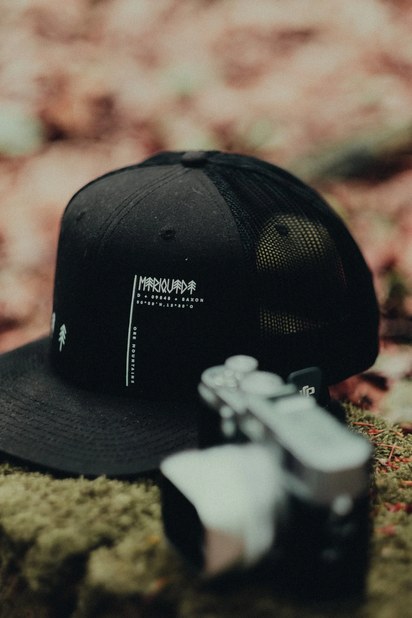 A black baseball cap with white text and a maple leaf logo, resting on a mossy surface. There is a smaller object in the foreground, possibly a camera or gadget.