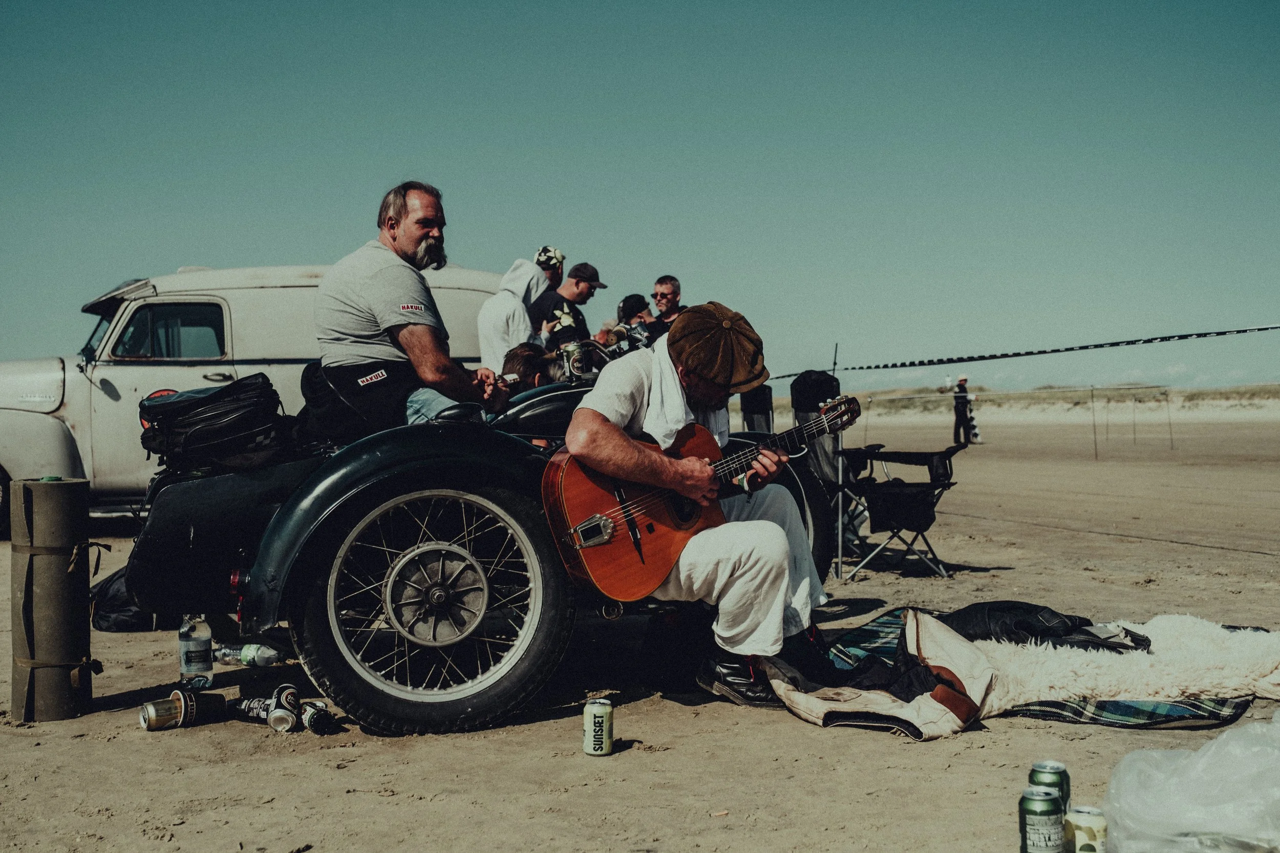 Group of people relaxing on a sandy beach with a vintage car, some sitting and others standing, one person playing an acoustic guitar, and others socializing under a clear sky.