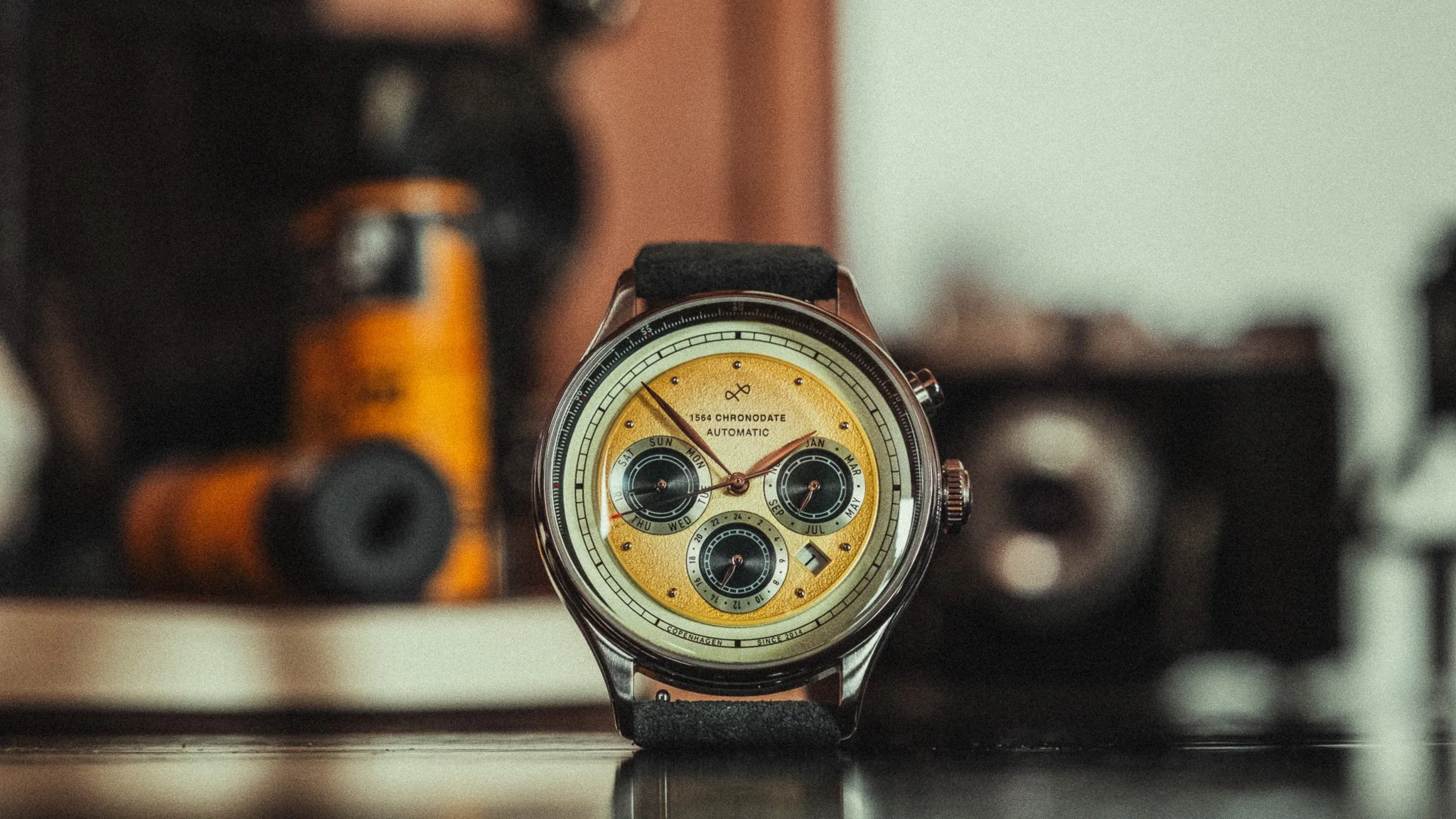 A close-up of a wristwatch with a yellow face, multiple dials, and a black strap, placed on a dark surface with blurred objects in the background.