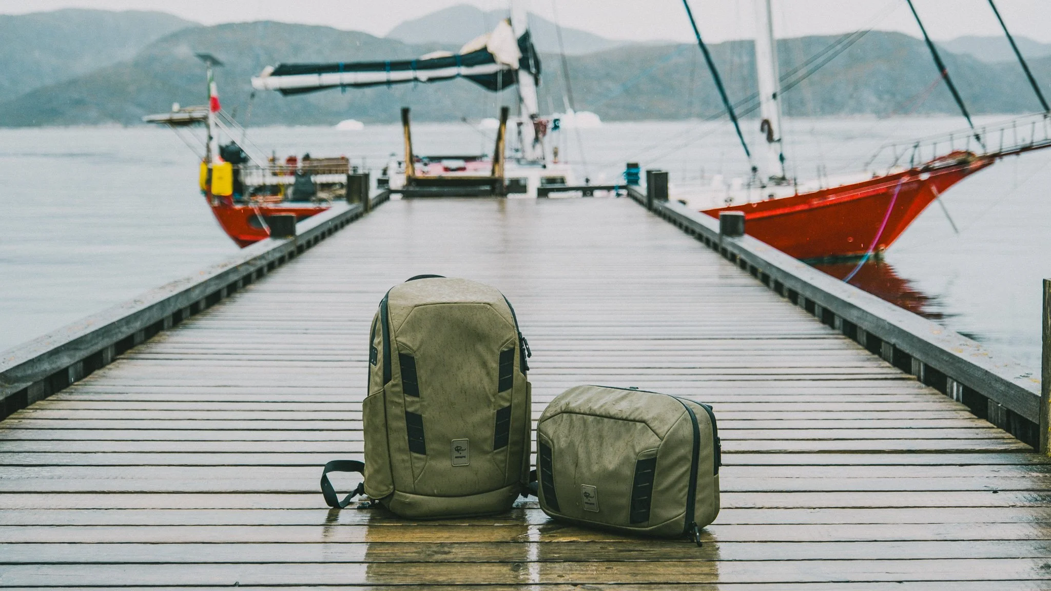 A view of a wooden dock with a backpack and a smaller bag on it, overlooking a calm body of water with a red sailboat docked nearby and mountains in the background.